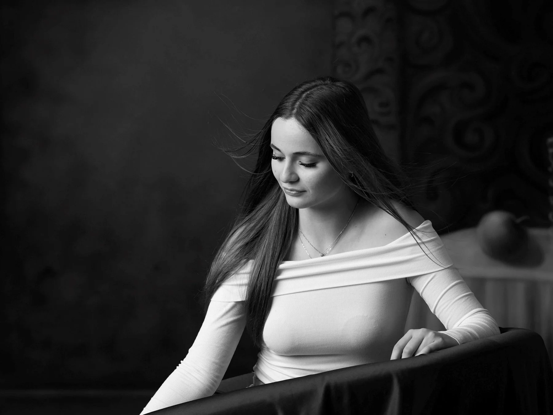 A young woman with long dark hair, wearing a light-colored off-the-shoulder top, looking down with her eyes closed, against a dark background with abstract patterns.