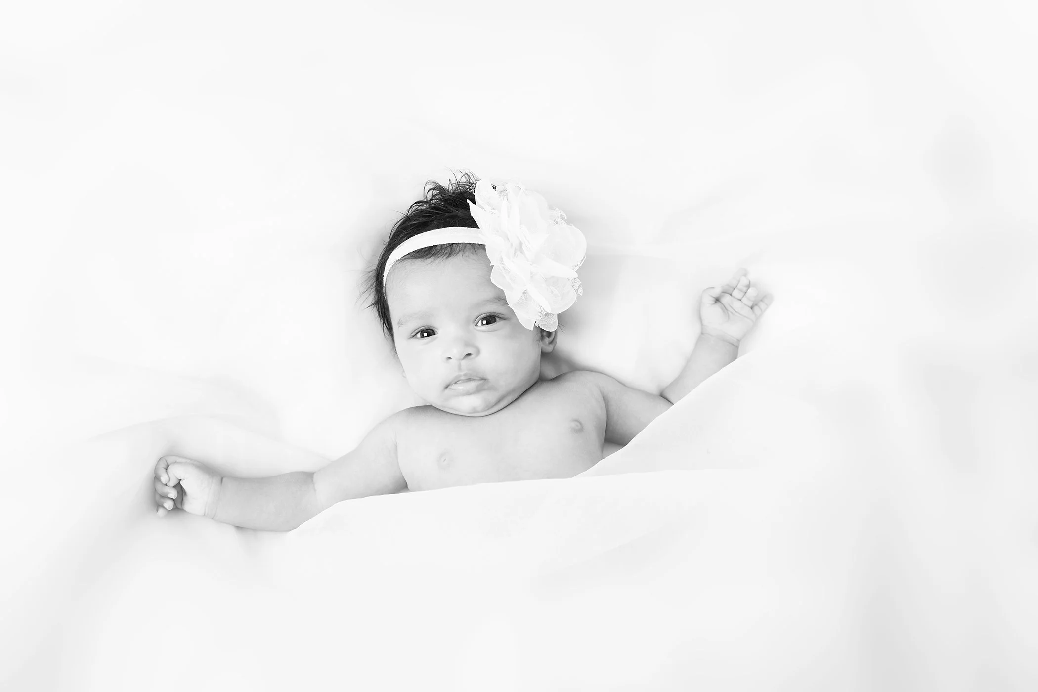 A baby girl with dark hair, wearing a large white bow headband, lying on a soft white surface, looking directly at the camera with one arm raised.