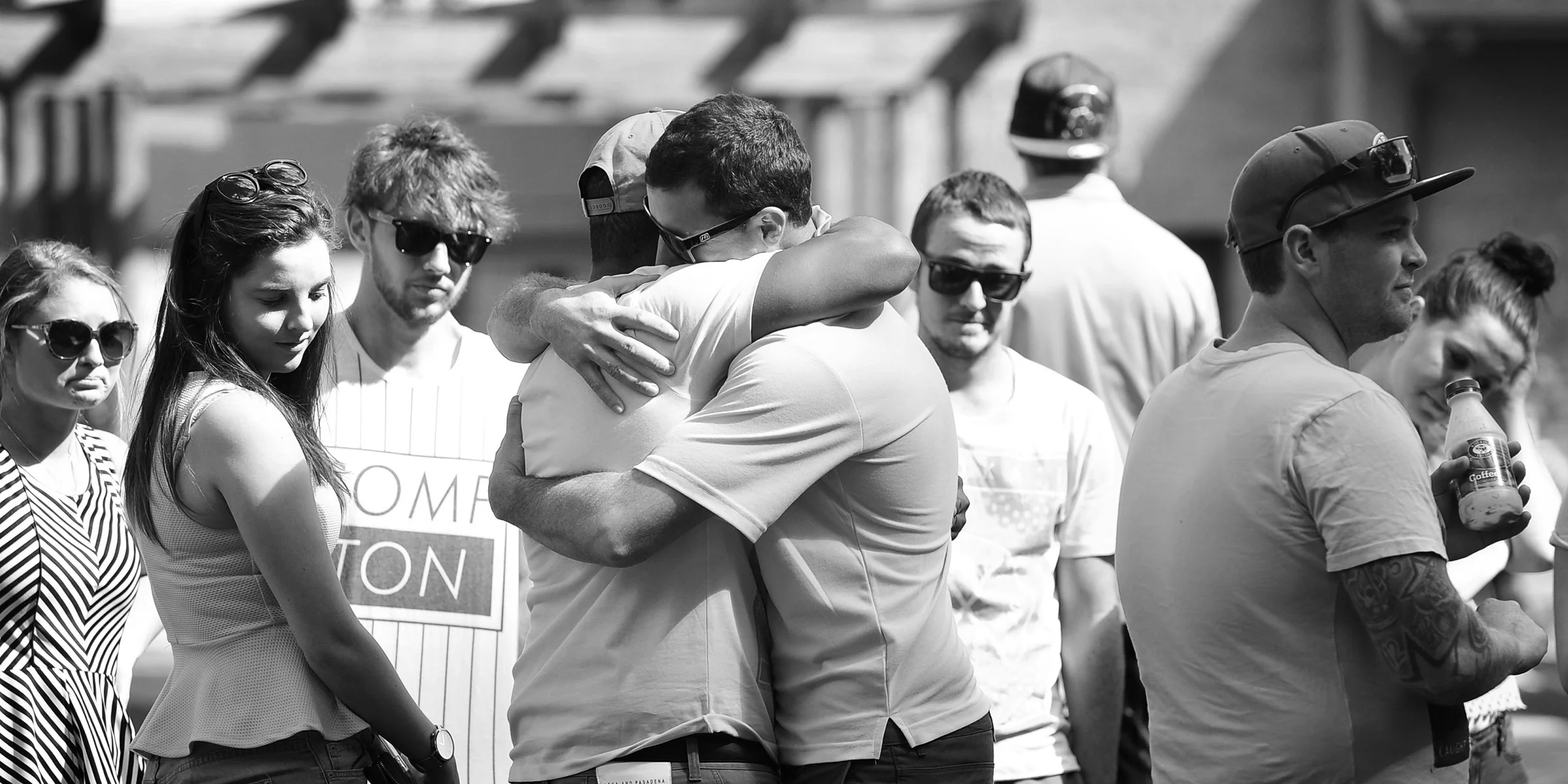 Two men are hugging each other in a crowd, with several women and men standing nearby, some wearing sunglasses and holding drinks, in an outdoor setting.