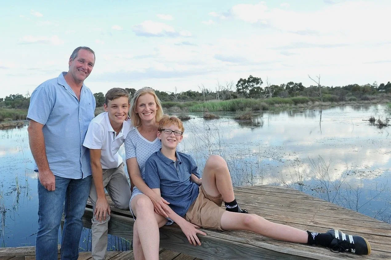 A family of five smiling on a wooden dock by a pond or lake, with trees and cloudy sky in the background.
