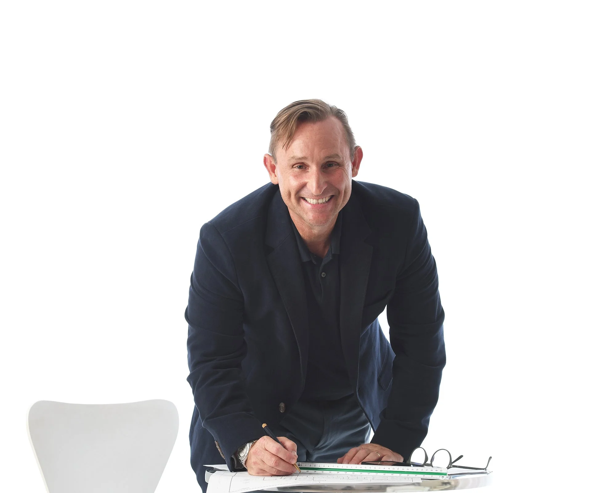 A smiling man in a dark suit leaning over a table writing with a pencil.