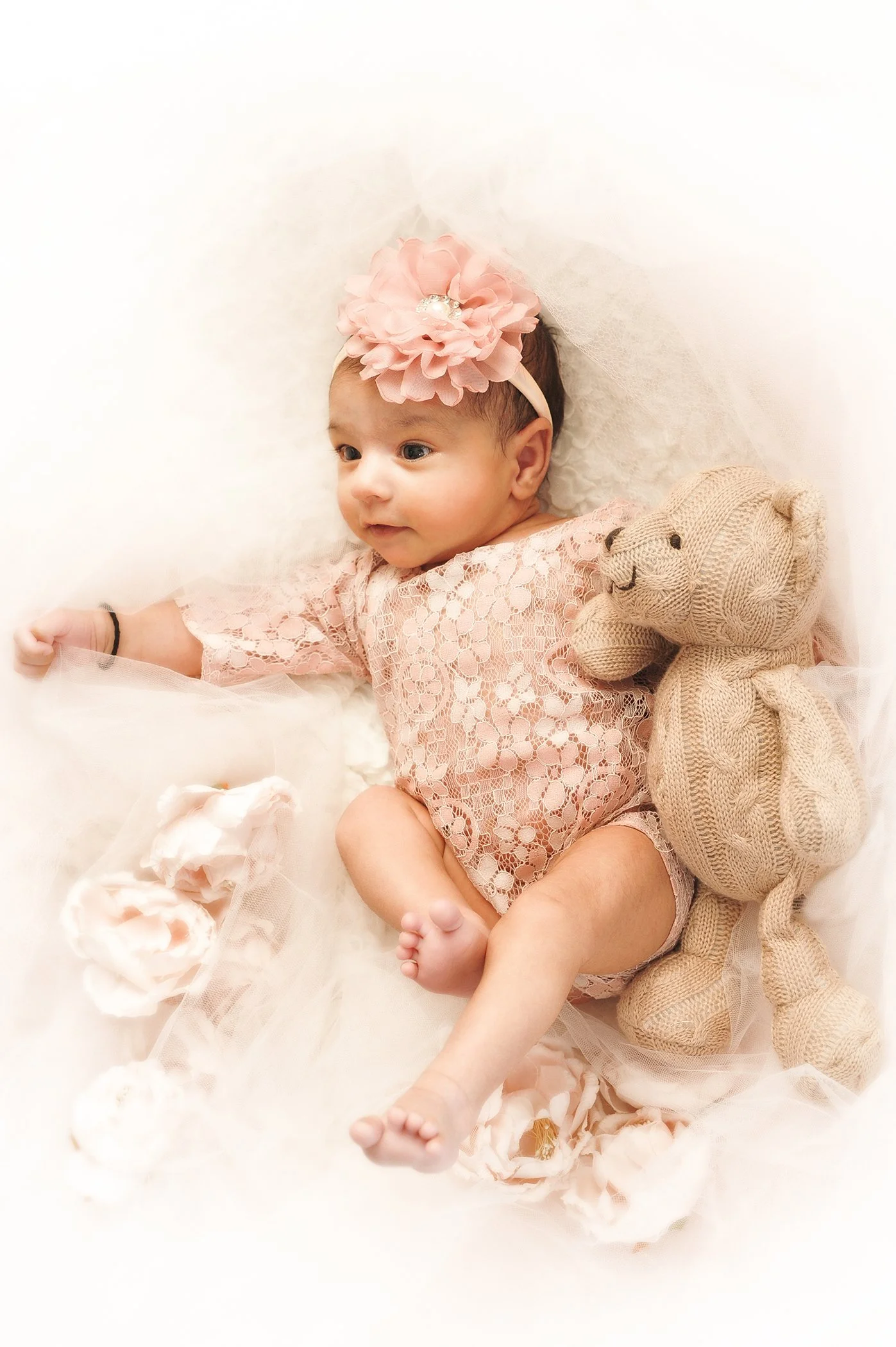 A baby girl wearing a pink lace dress and a large pink flower headband, lying on a soft, creamy surface with a beige teddy bear and peach-colored roses nearby.