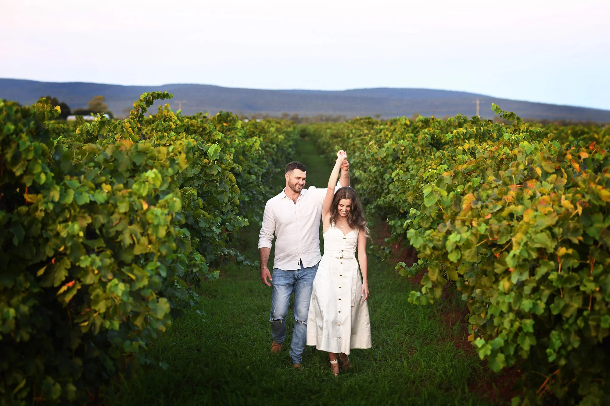 A young couple walking through a vineyard, with the woman holding the man's hand raised in the air, both smiling, greenery on either side, with distant hills and a clear sky in the background.