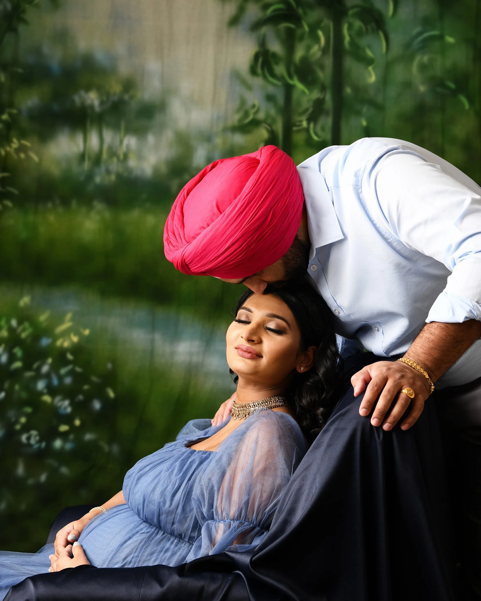 A man wearing a red turban and white shirt gently kissing a woman with closed eyes, dressed in a traditional outfit, while holding her in his arms in a serene outdoor setting with blurred greenery and a metal railing in the background.