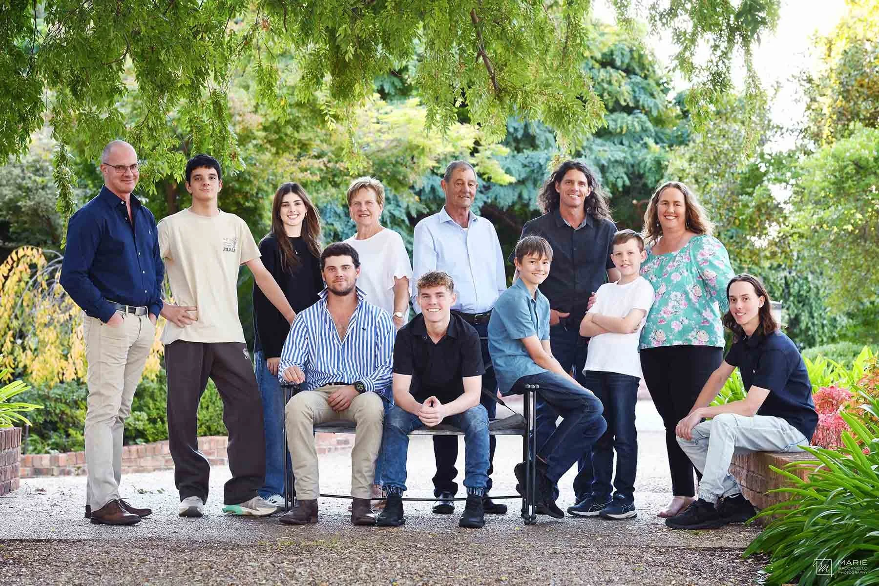 A diverse group of fifteen people, including adults and children, posing outdoors in a garden with lush green trees and plants. They are smiling and arranged in a semicircle, some standing and some seated on chairs, with a brick pathway and vibrant f