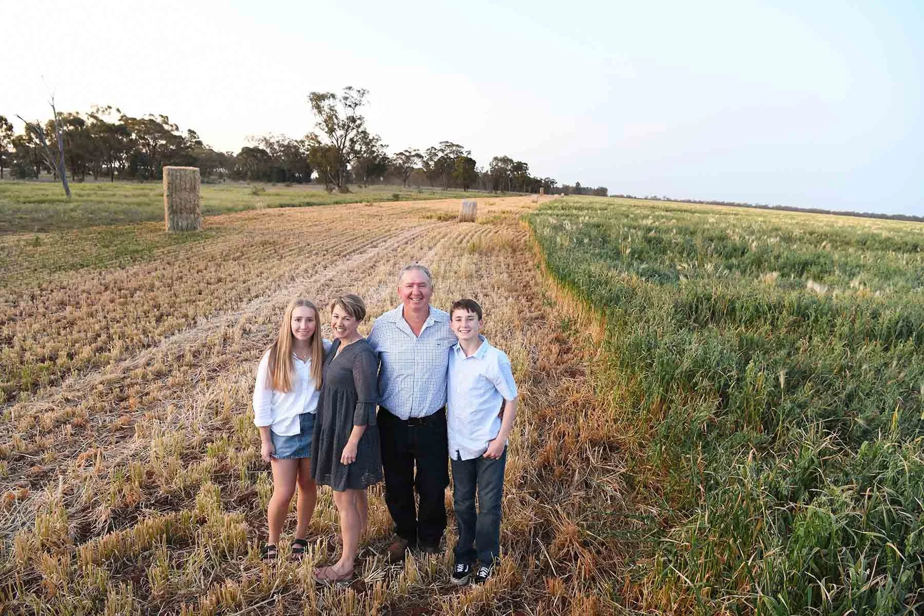 Family of four standing in a rural field divided into a harvested area on the left and a green unharvested crop on the right, with trees and hay bales in the background.
