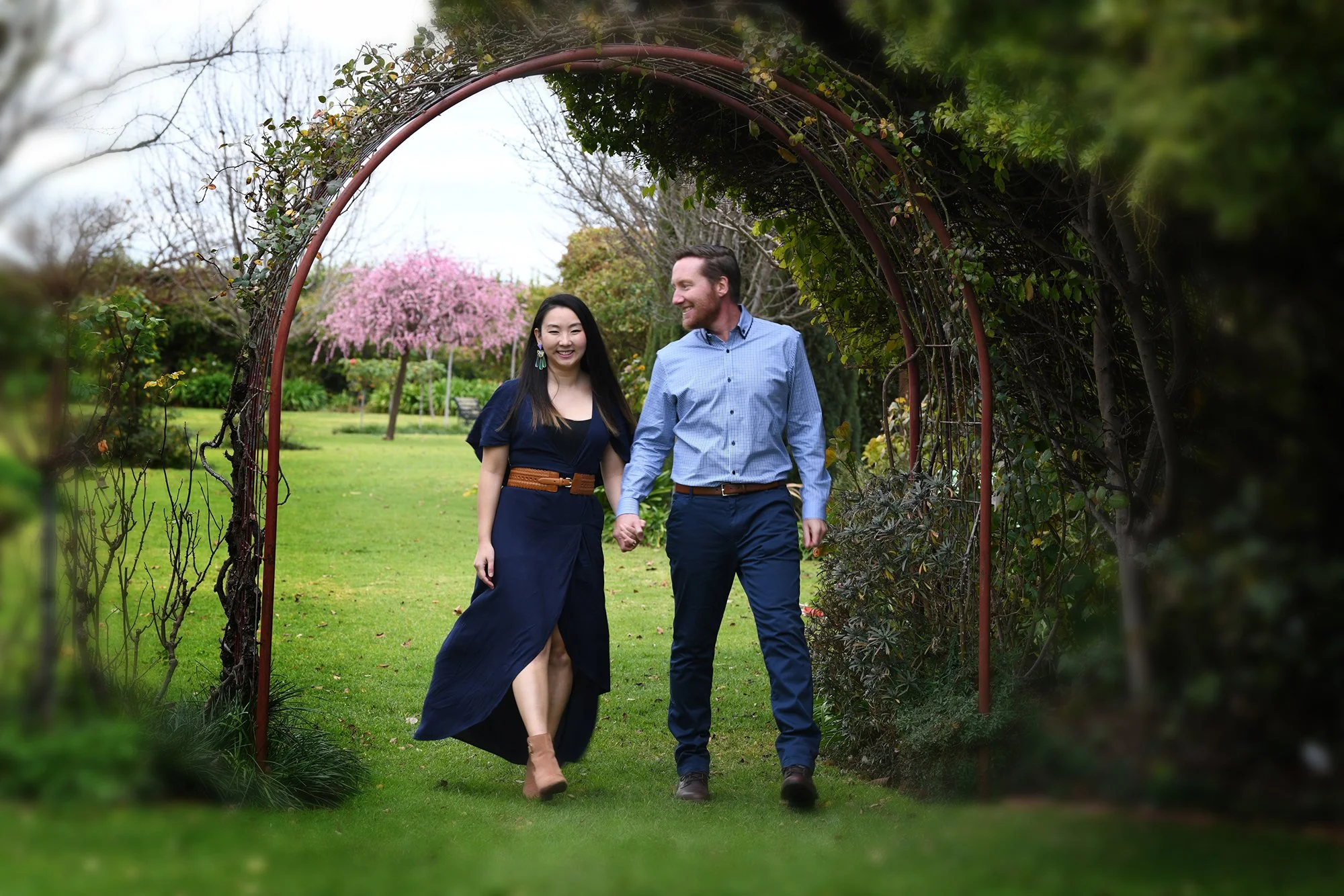 A smiling couple holding hands and walking through a garden archway covered with vines, with trees and pink flowering trees in the background.