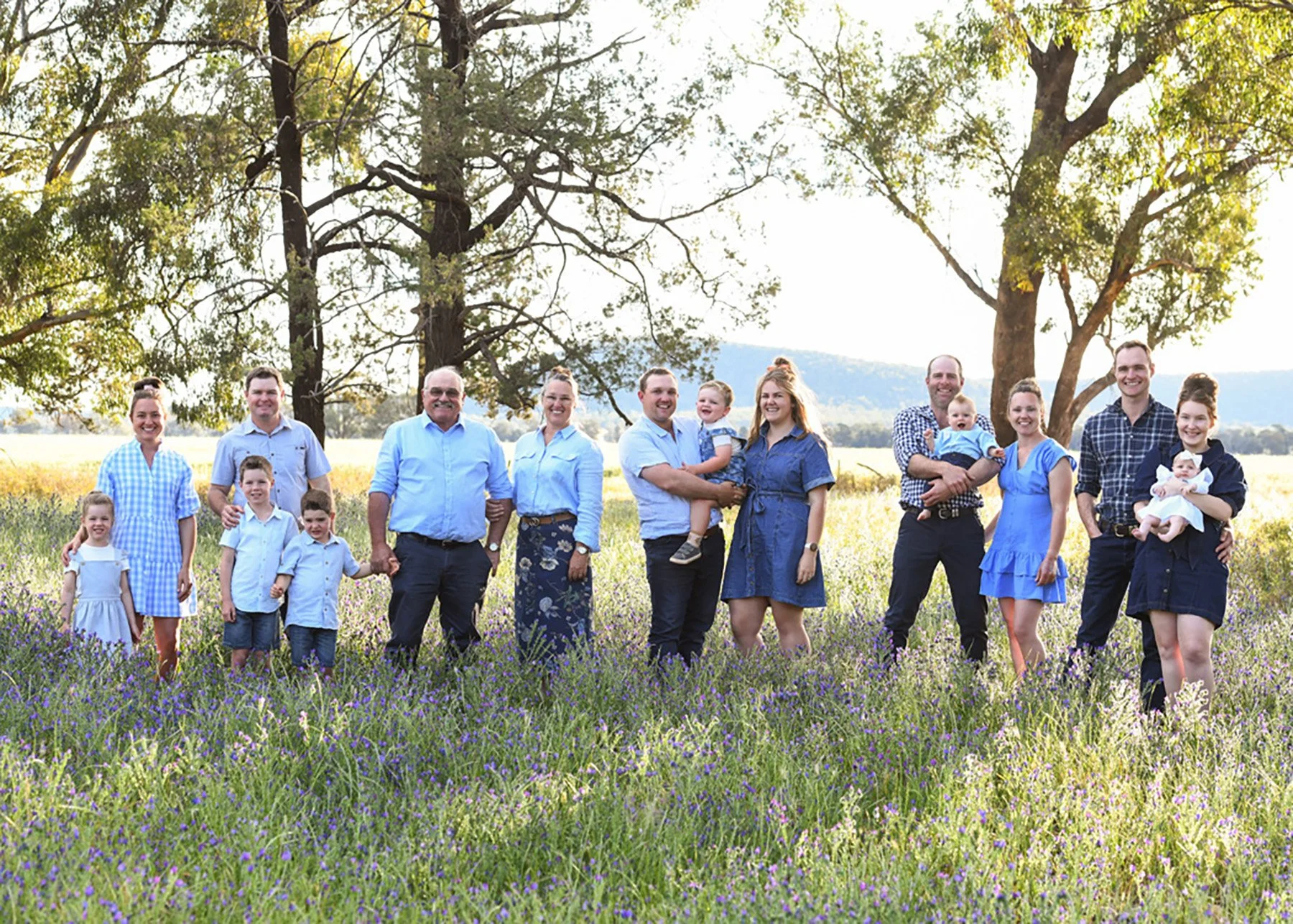 A large family standing in a field of purple flowers with trees and hills in the background, smiling at the camera on a sunny day.