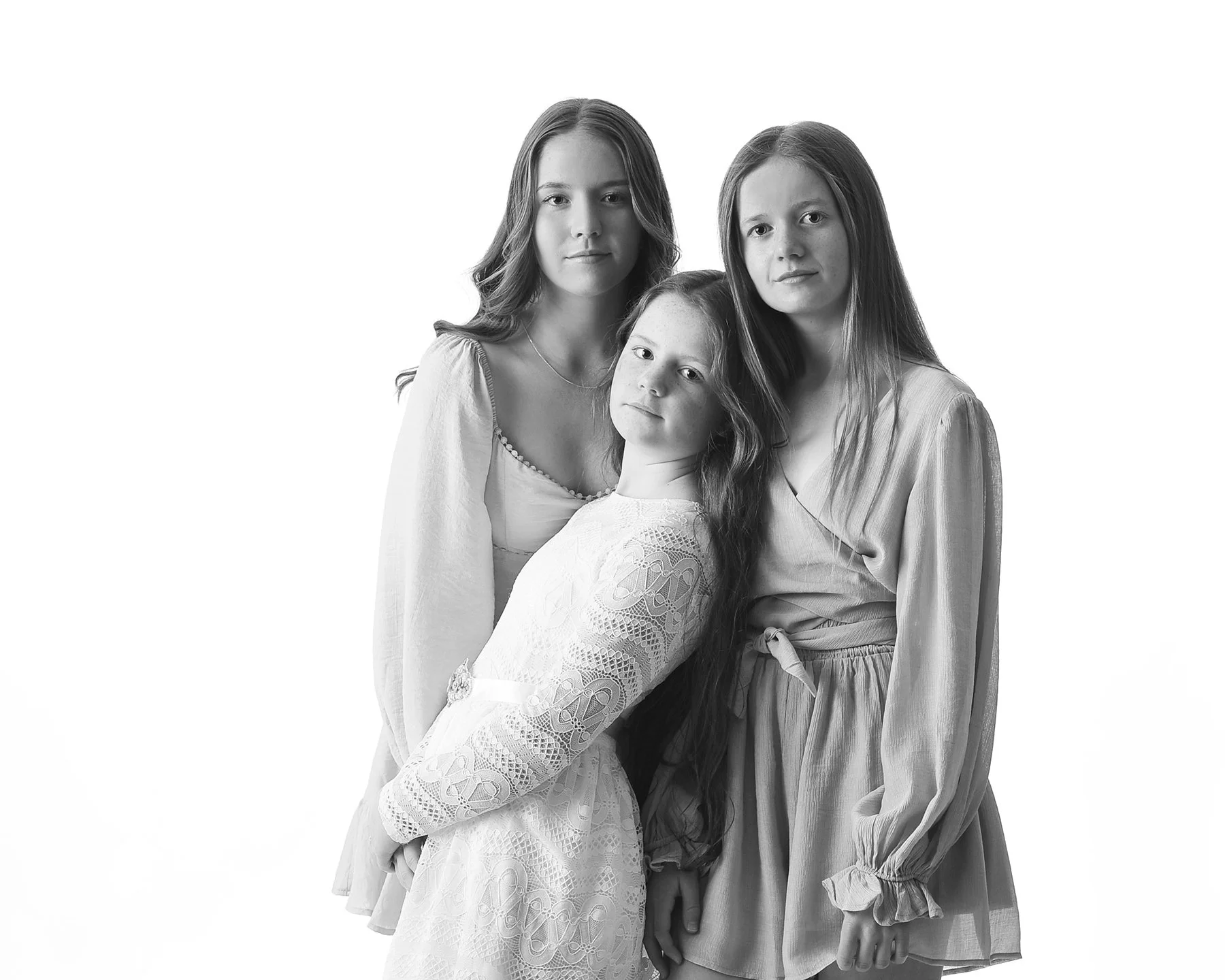 Black and white photo of three young girls standing close together, looking at the camera with neutral expressions, against a plain light background.