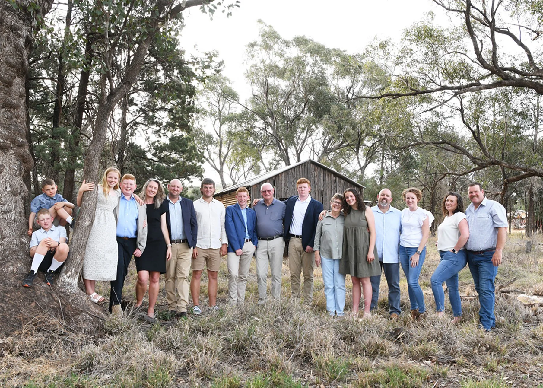 A large group of people, including children and adults, standing outdoors in front of a rustic wooden shed and tall trees, smiling and posing for a group photo.