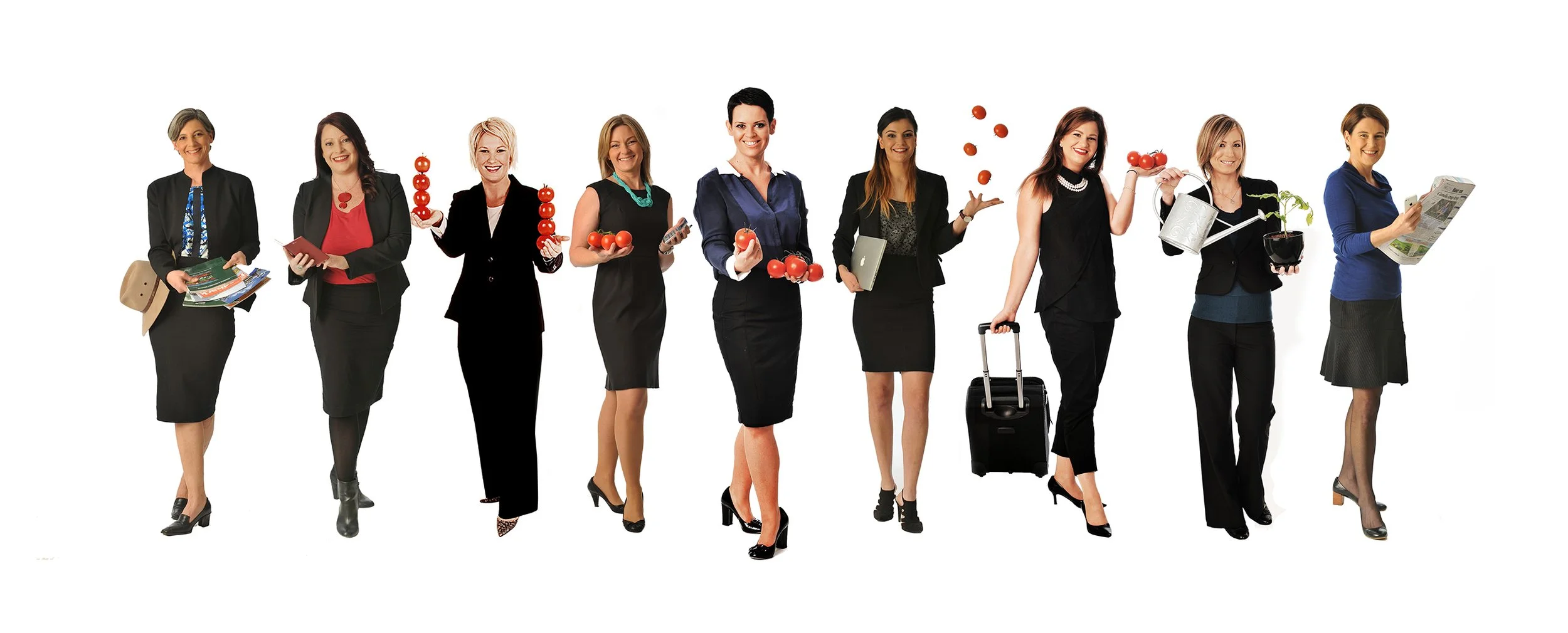A group of ten professional women standing in a line, each holding or interacting with various objects such as books, tomatoes, a laptop, a watering can with a plant, and a newspaper, dressed in business attire, against a white backdrop.