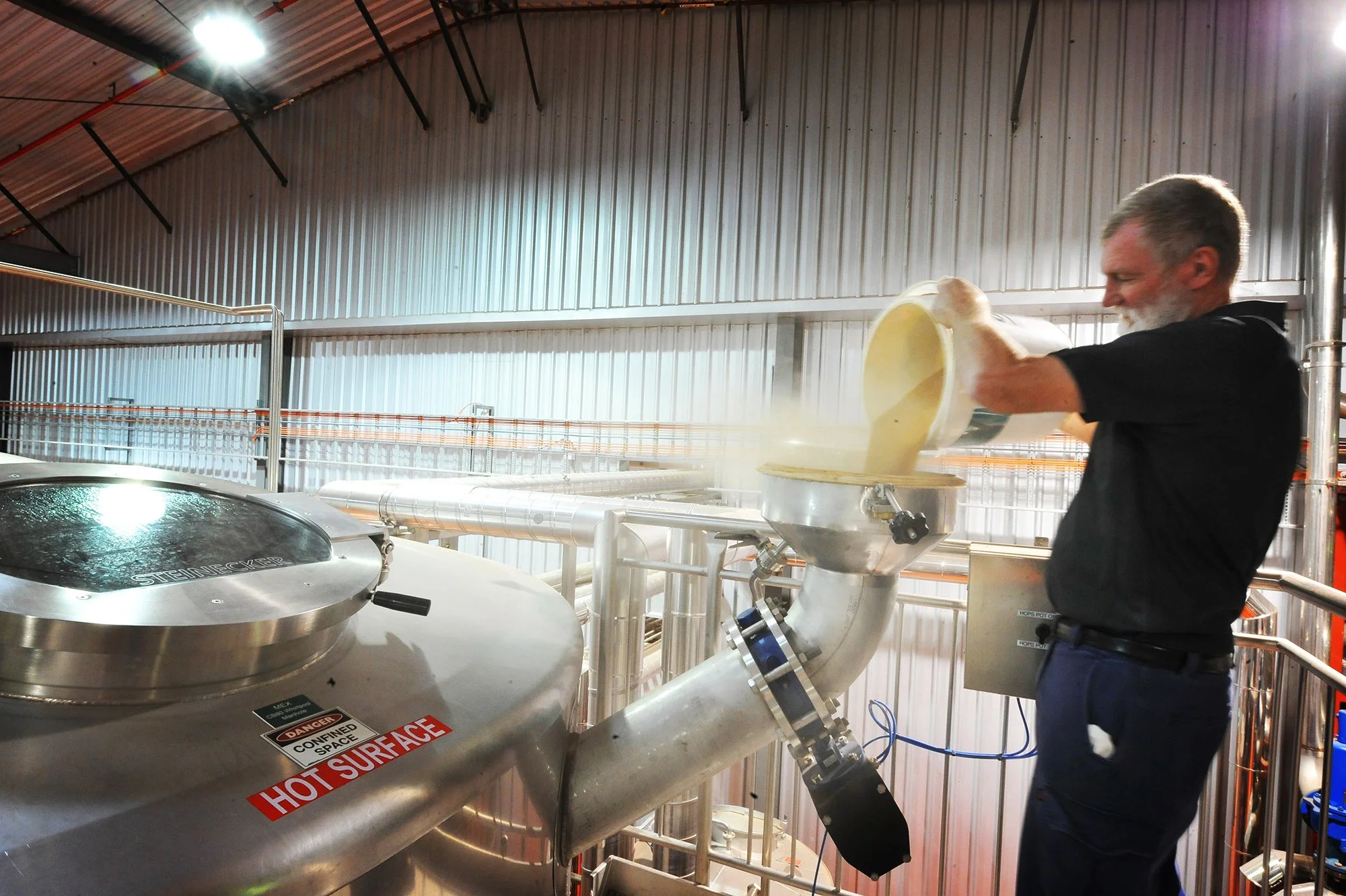 Man pouring liquid into large stainless steel tank in industrial setting with warning labels.