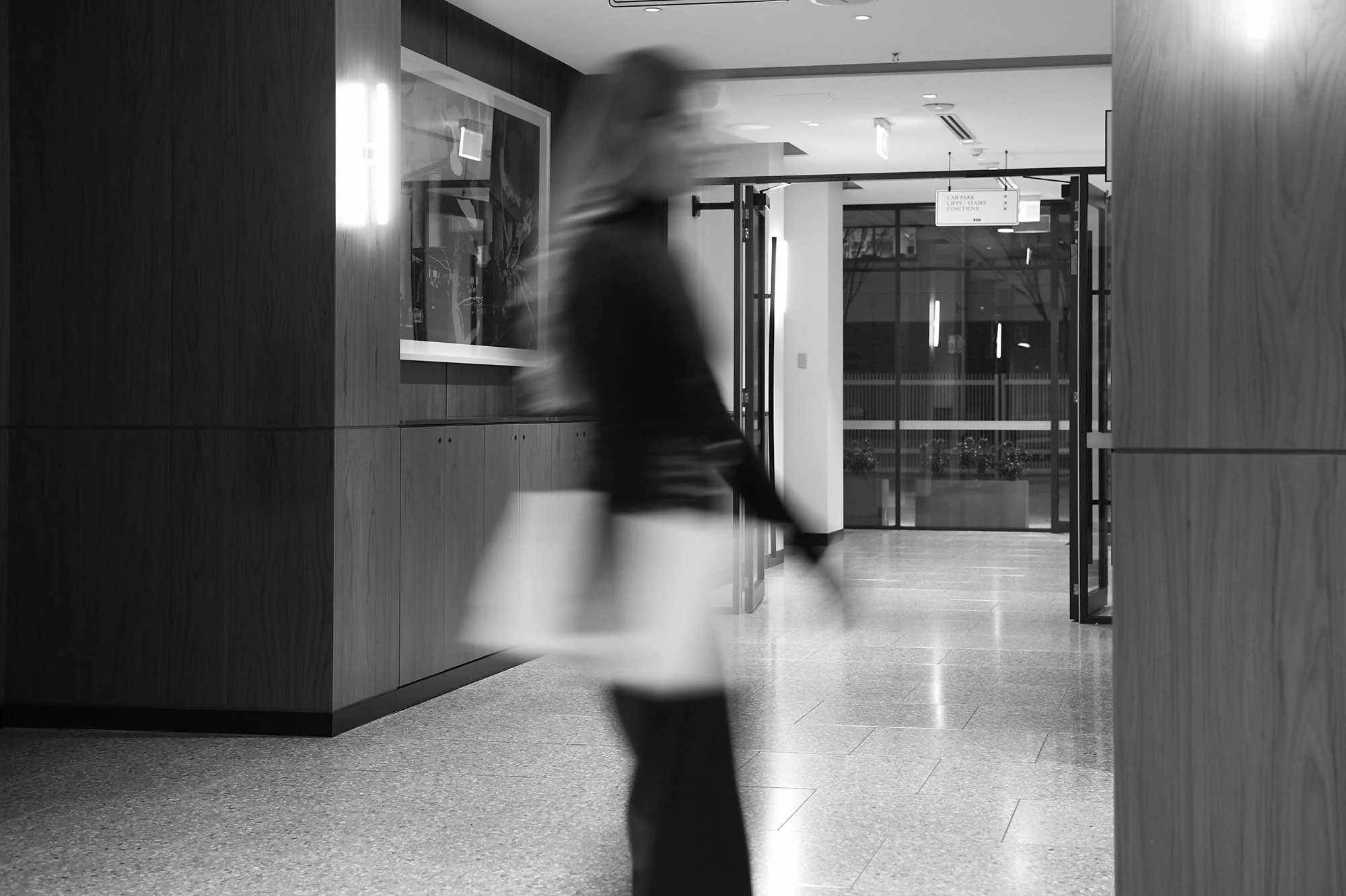 Blurred person walking through an indoor lobby area with large glass doors in the background, black and white photograph.