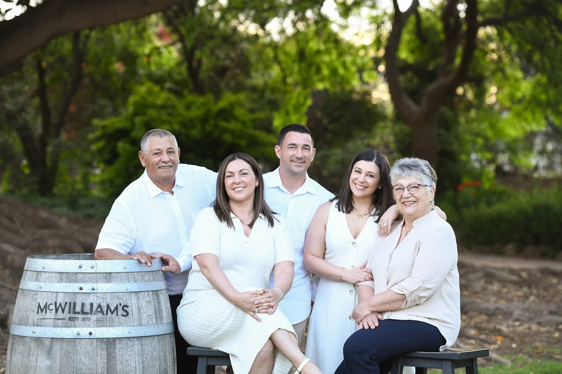 A group of six people standing outdoors in a park-like setting, smiling, with trees in the background. They are dressed in light-colored clothing, and there is a wooden barrel labeled 'McWilliam's' in the foreground.