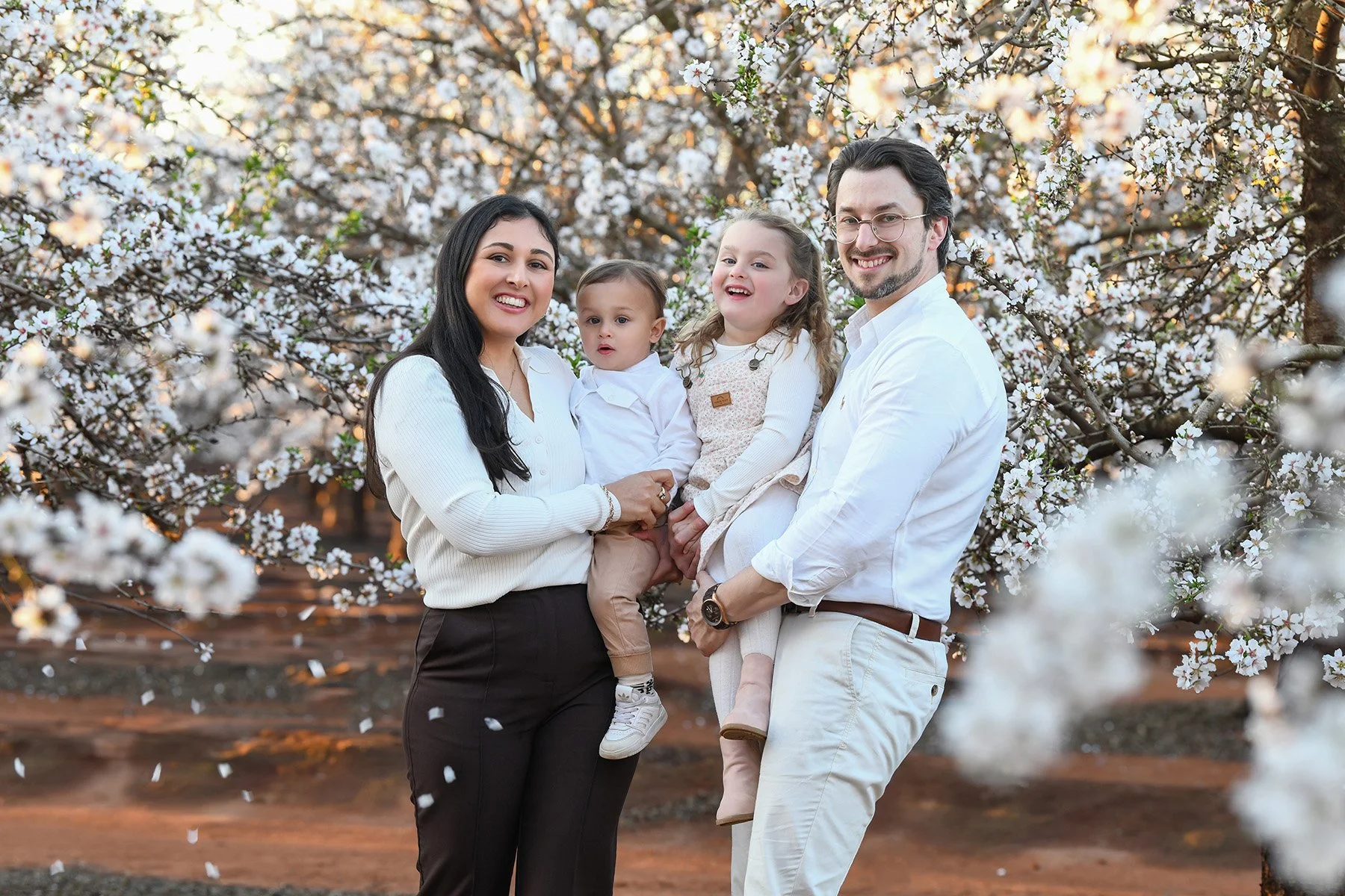 Happy family of four in springtime, standing outdoors among blooming cherry blossom trees. The mother and father are holding their children, a boy and a girl, smiling at the camera.