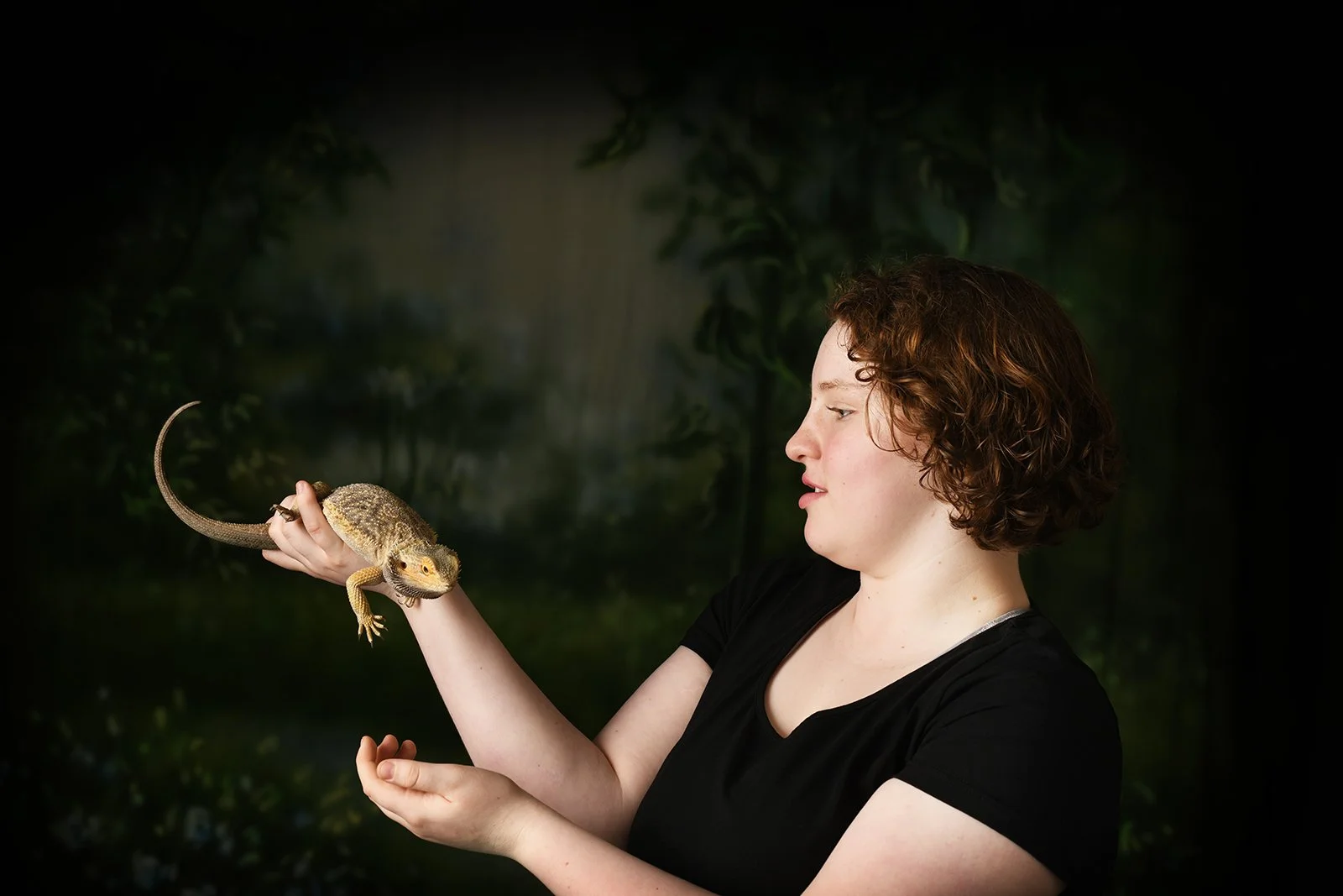 Young woman with curly brown hair holding a small lizard in her hand, with a dark green, blurred background.
