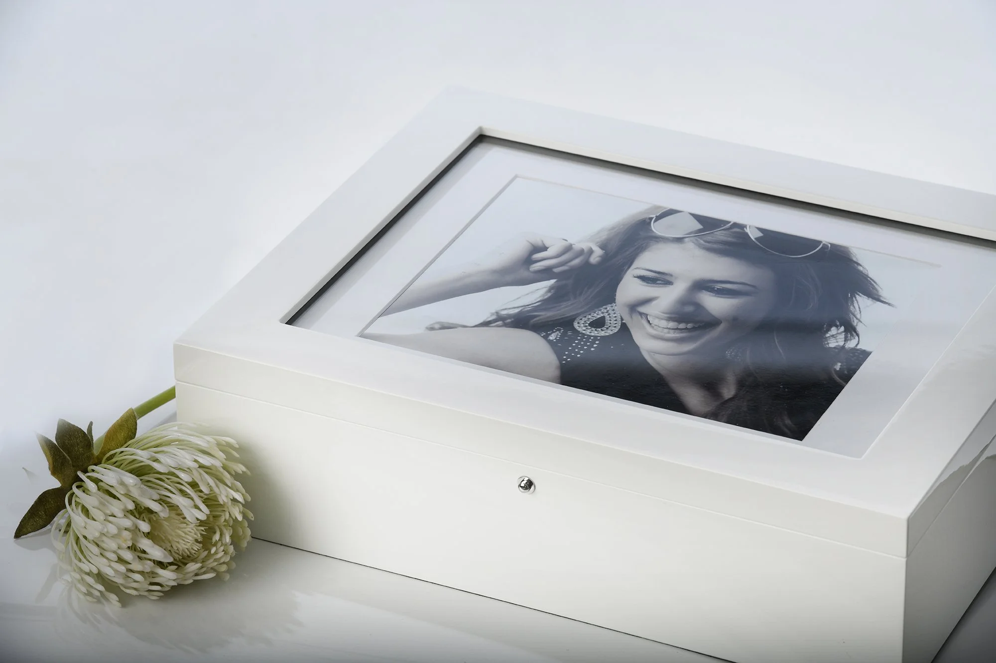 A framed black and white photograph of a smiling woman with sunglasses is inside a white box with a glass top. A white flower with green leaves lies beside the box.