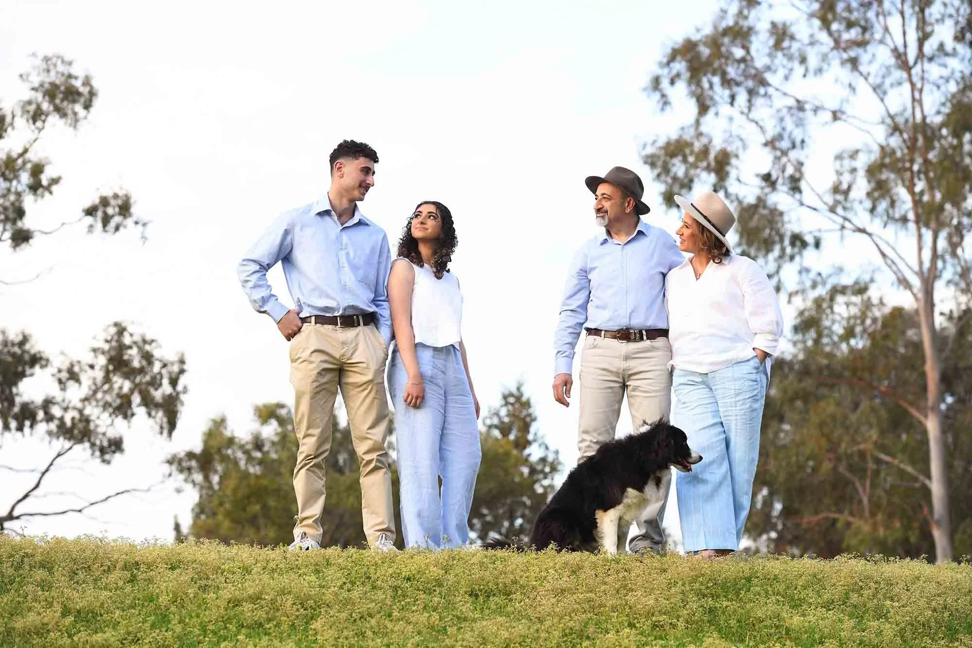 Four people and a dog standing outdoors on grass, with trees in the background. The group appears to be enjoying a casual moment together.