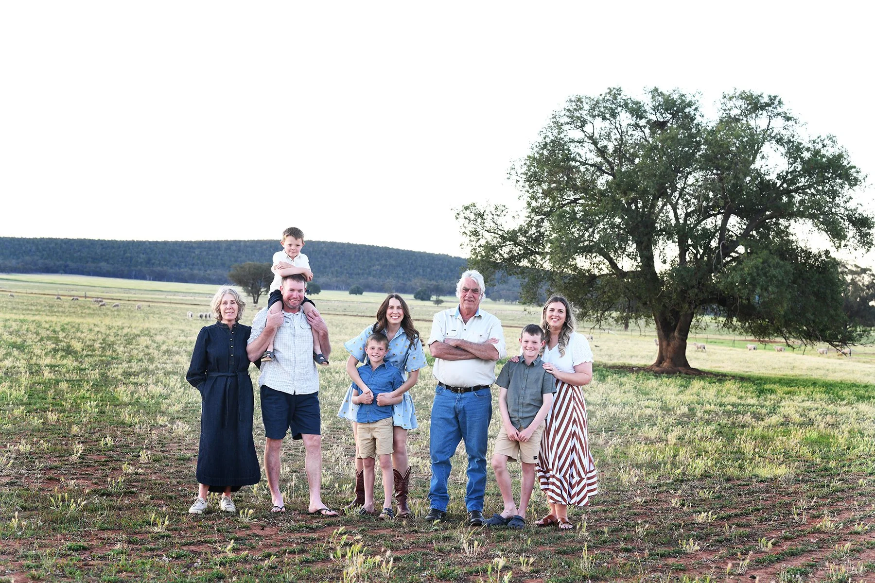 A family of eight standing in a field with a large tree and cows in the background, during daytime.