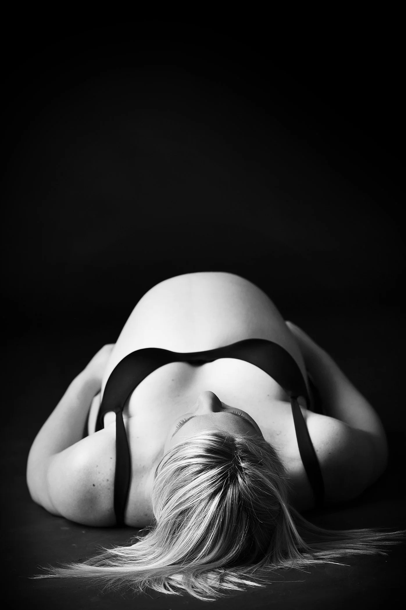 A black and white photo of a woman lying on her back with her head tilted to the side and her long hair spread on the surface. She is wearing a black bra and appears to be looking upward.