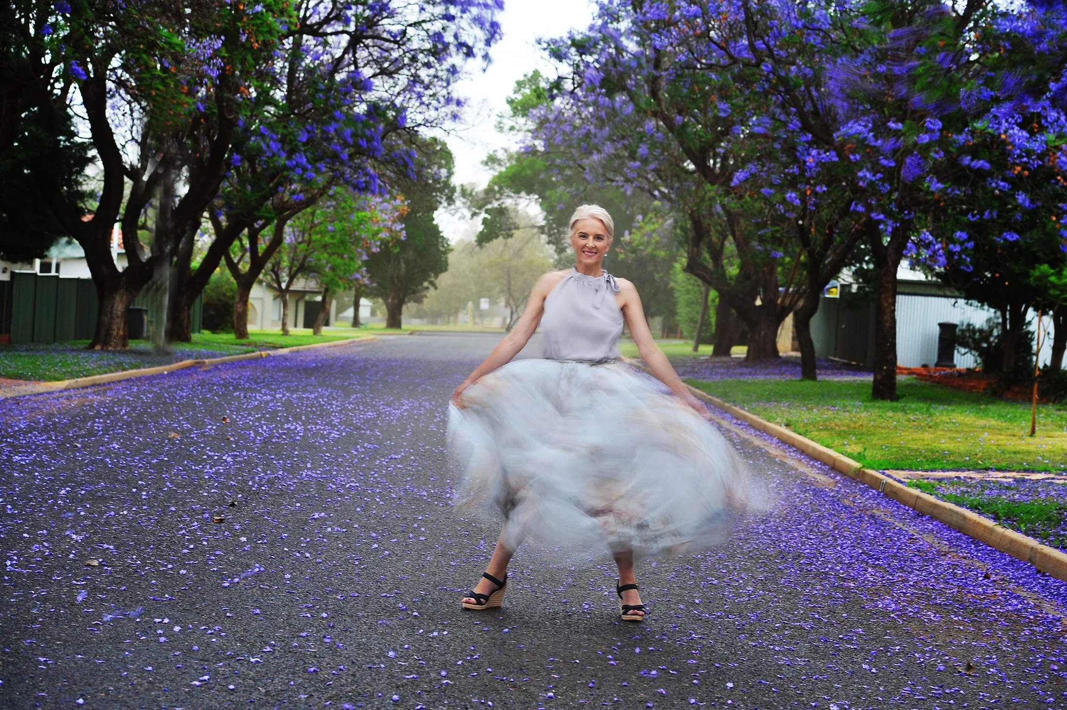 A woman in a light-colored dress with a tulle skirt and high-heeled sandals posing on a paved street lined with trees that have purple flowers, with fallen purple petals on the ground.
