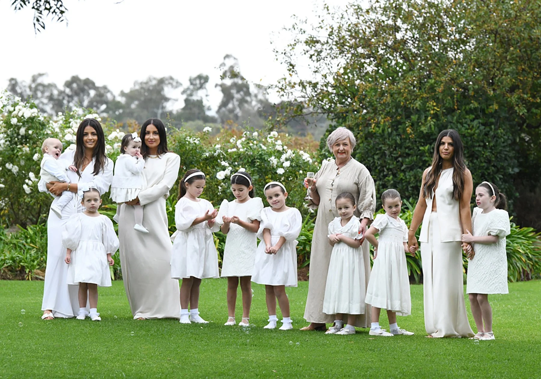 A group of women and young girls dressed in white standing on a lush green lawn with blooming white flowers and trees in the background