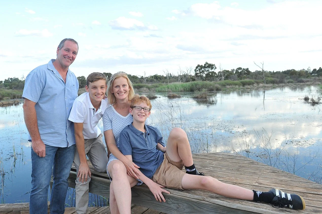 A family of five standing and sitting on a wooden dock by a calm body of water with trees and cloudy sky in the background.