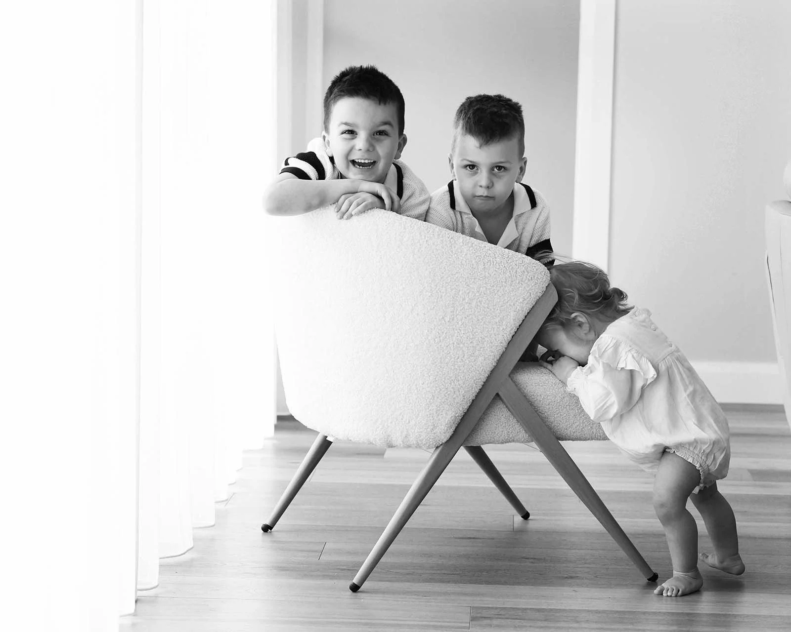 Two older boys leaning over a modern armchair, watching a young girl leaning into the chair, with one boy smiling and the other with a serious expression.