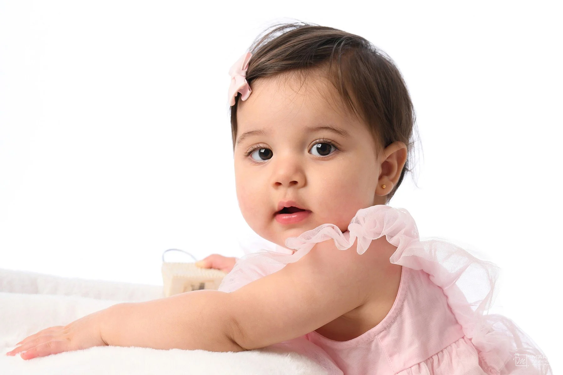 A young girl with dark hair, big eyes, and a pink bow in her hair, lying on her stomach on a white surface, wearing a pink dress with ruffled sleeves, looking at the camera with her mouth slightly open.