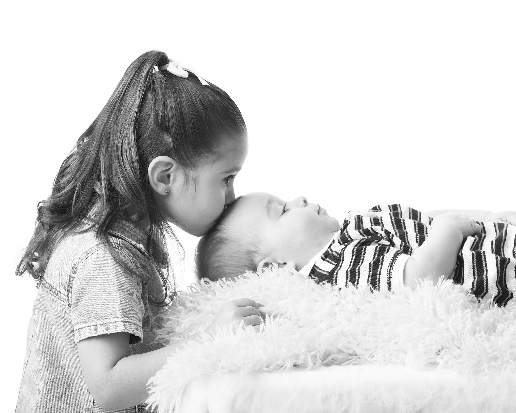 A young girl with long hair and a bow, face close to a baby lying on a soft surface, both engaging in a tender moment.