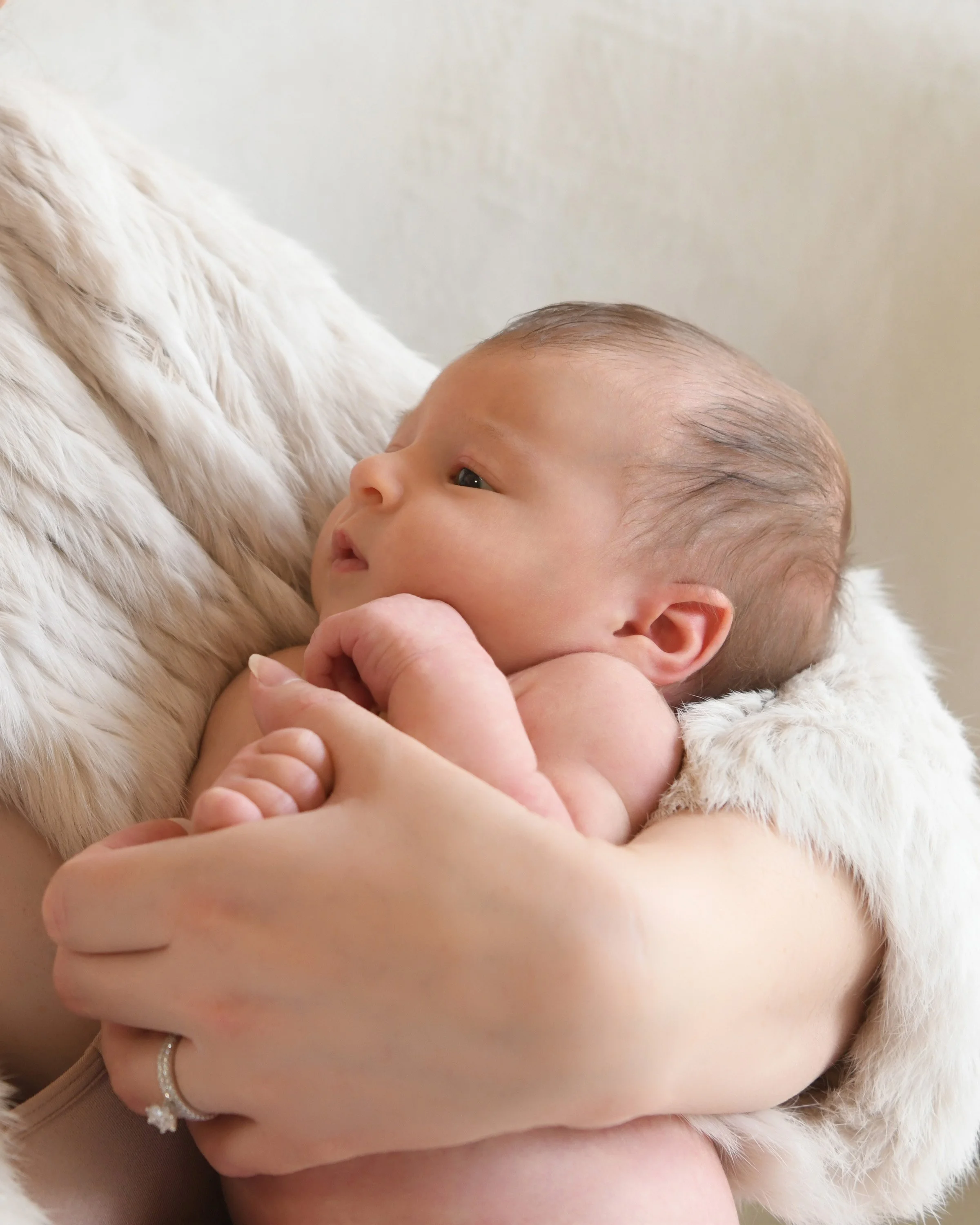 Close-up of a newborn baby held in someone's arms with soft, white fur on their clothing, and a ring on their finger.