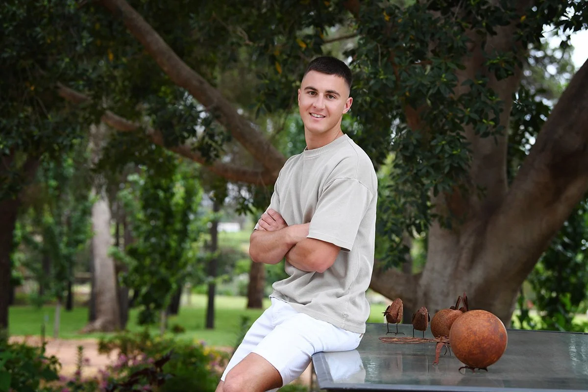 A young man with short dark hair, wearing a beige t-shirt and white shorts, sitting on a table outdoors in a park with trees in the background. There are decorative metallic sculptures and spherical objects on the table next to him.