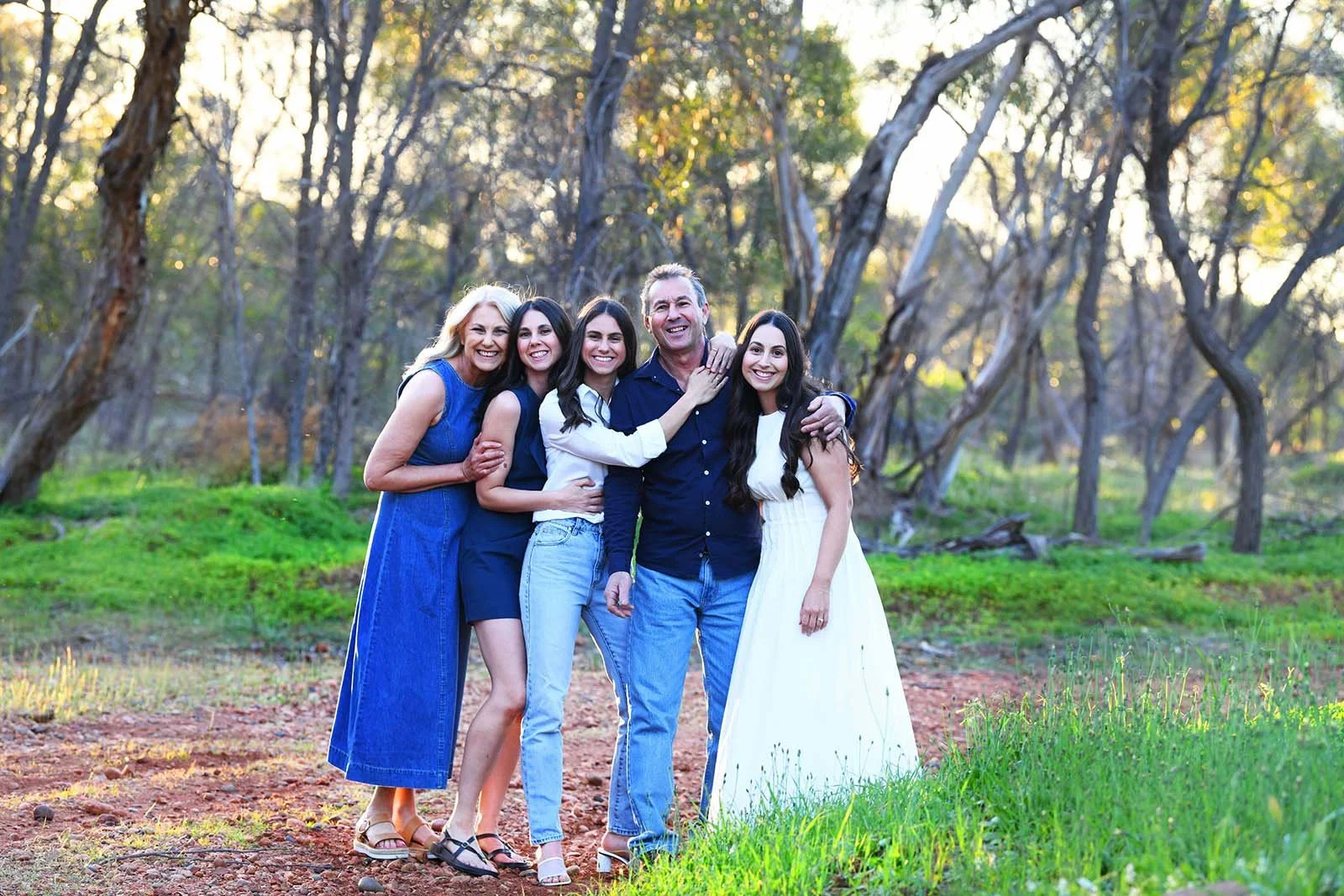 A family of five standing outdoors in a wooded area with green grass and trees, smiling and embracing each other.