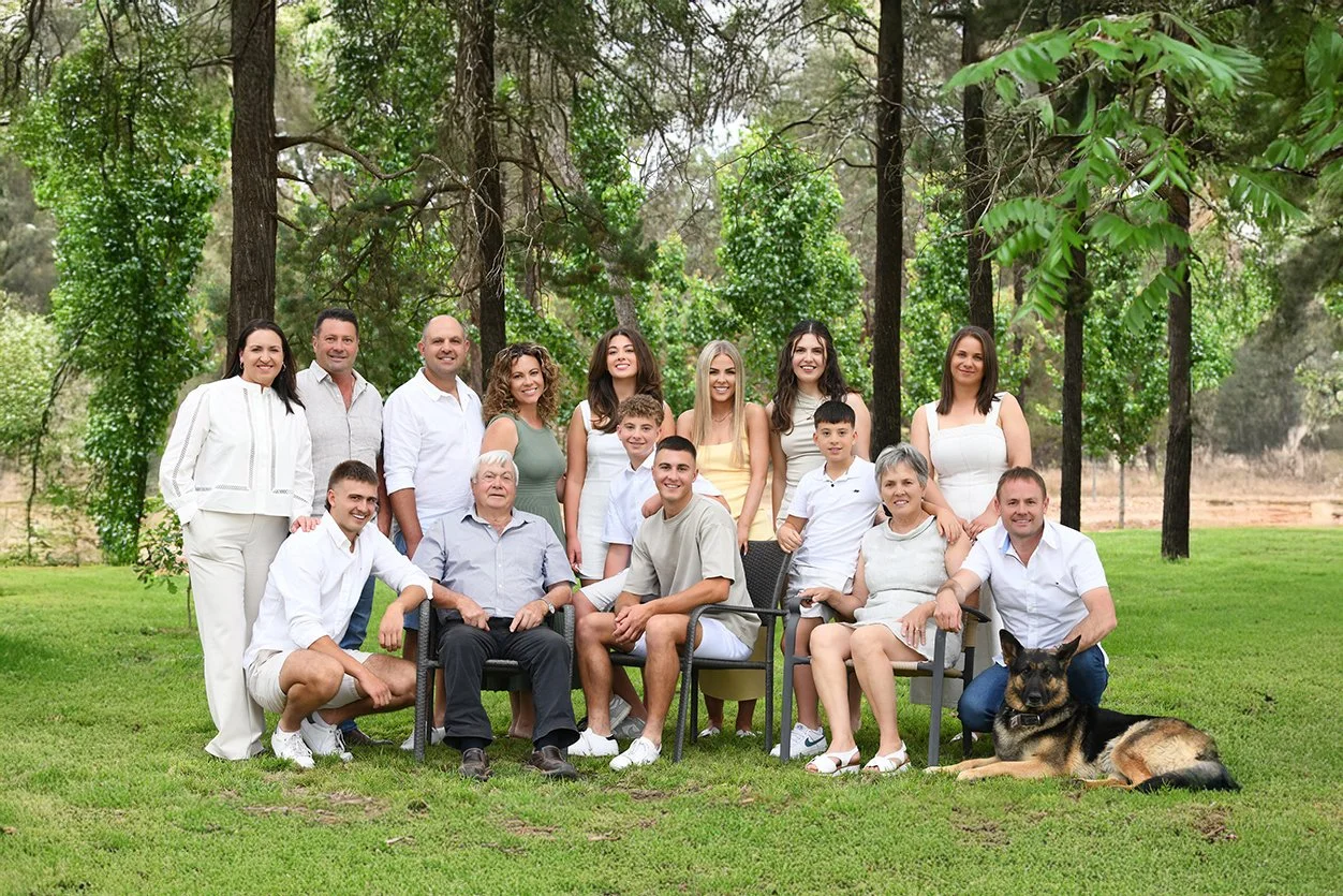 Large family group photo outdoors in a park, with family members of various ages, including children, adults, and seniors, sitting and standing on a grassy area among trees, with two dogs laying on the grass in front.