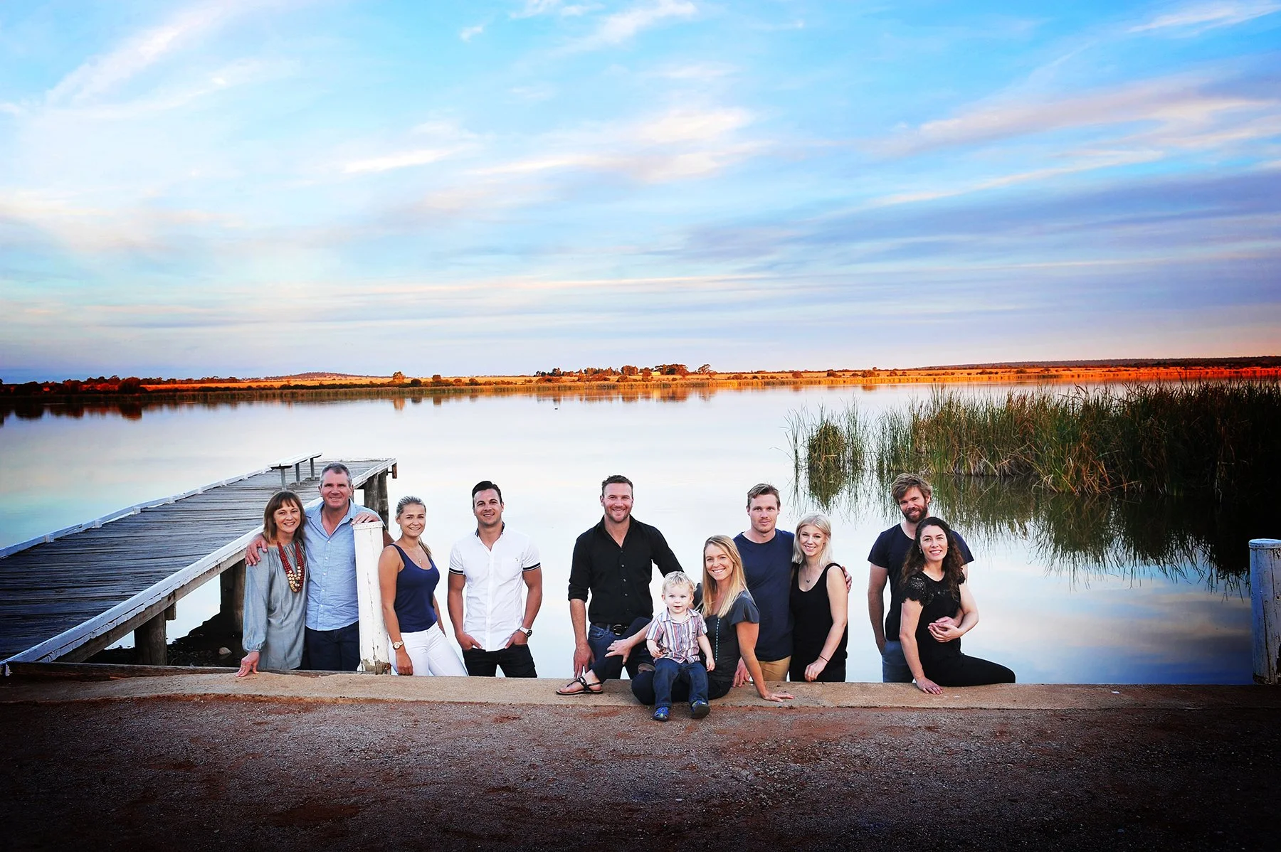 A group of ten people, including adults and children, standing on a dock by a calm lake during sunset, with tall grasses and a colorful sky in the background.