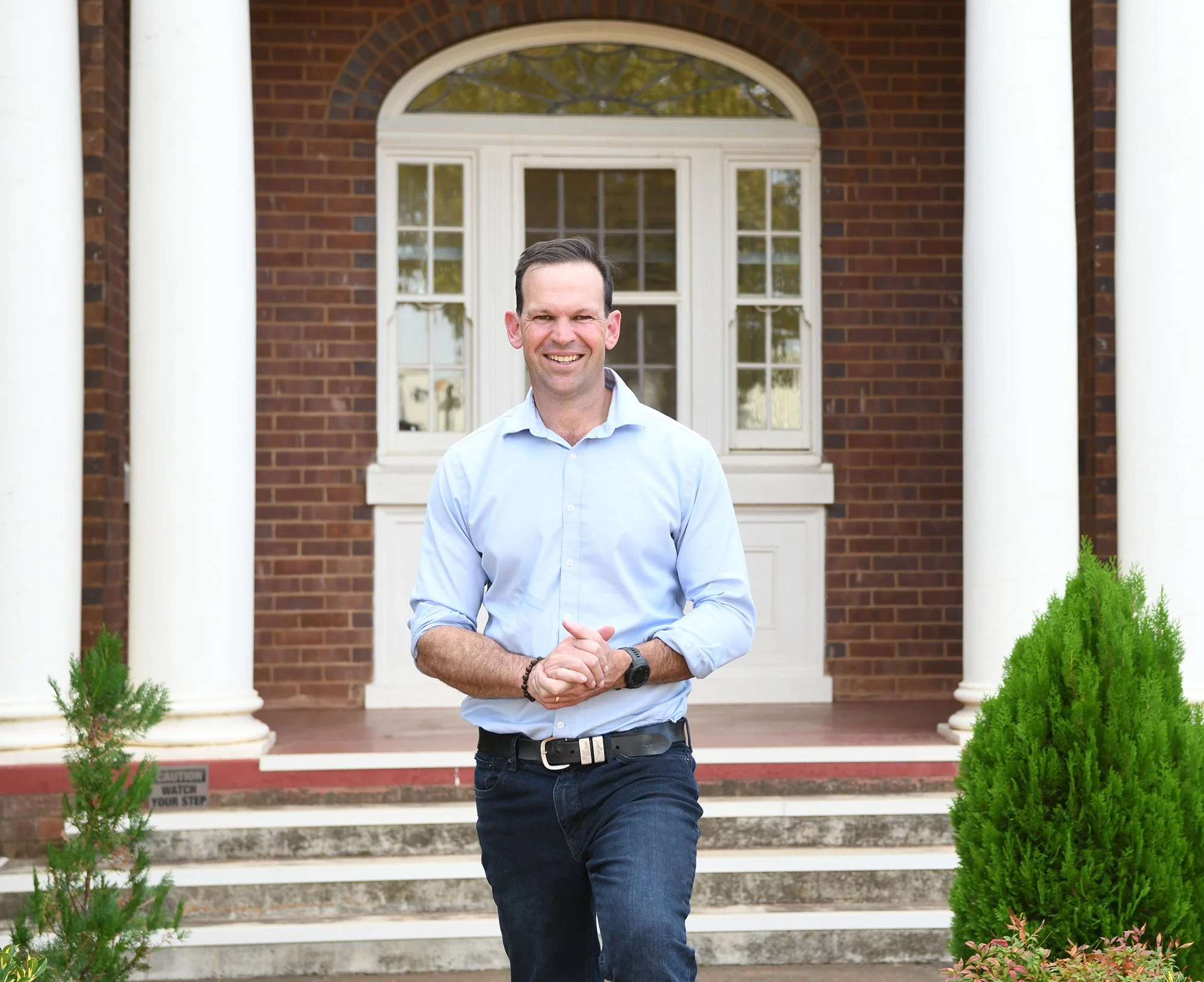 A man in a light blue shirt, black belt, and dark blue jeans standing in front of a brick house with white colonial columns and a large window.