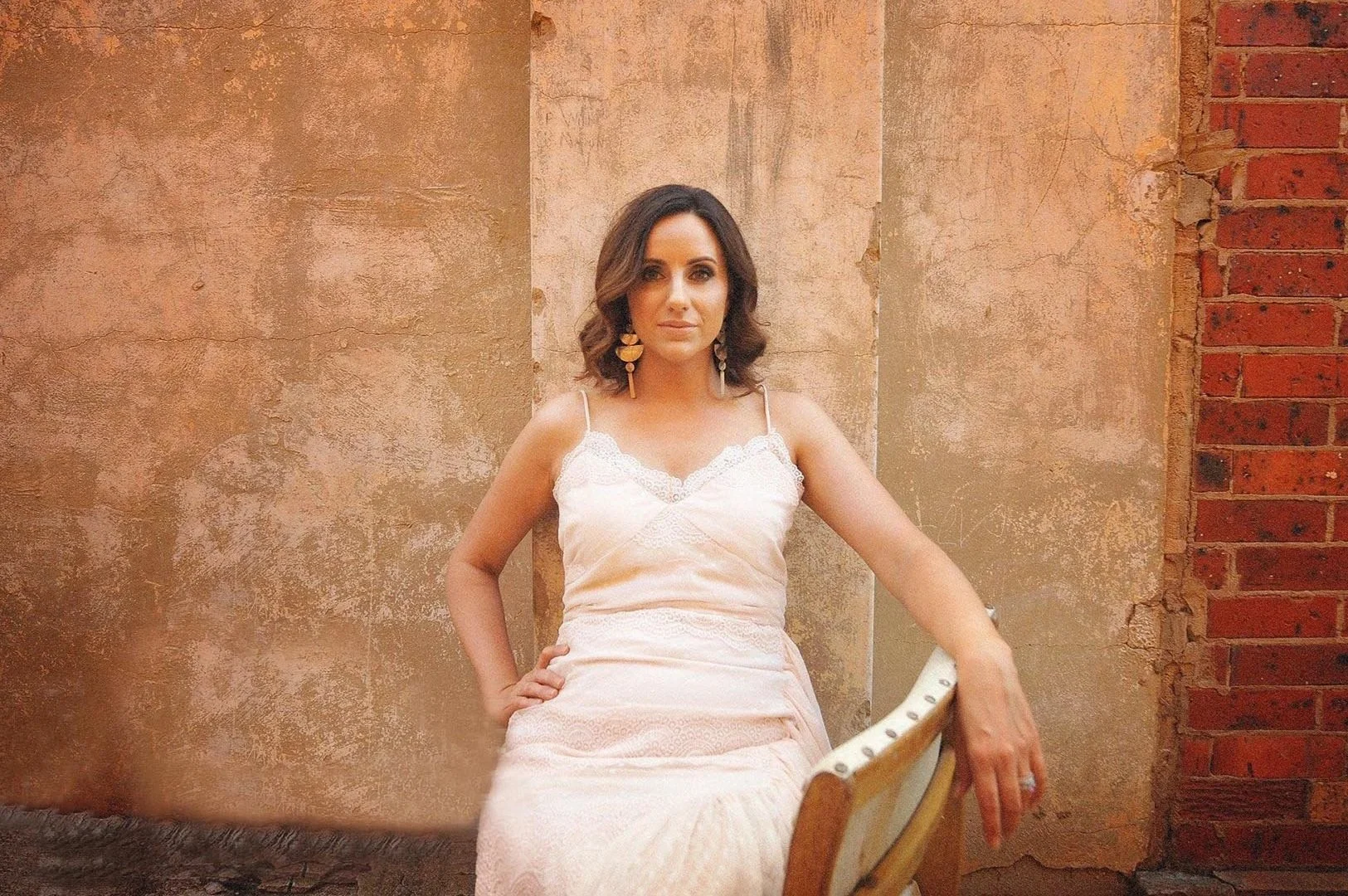 Woman in a white dress sitting on a chair against a textured beige wall, with a red brick wall on the right.