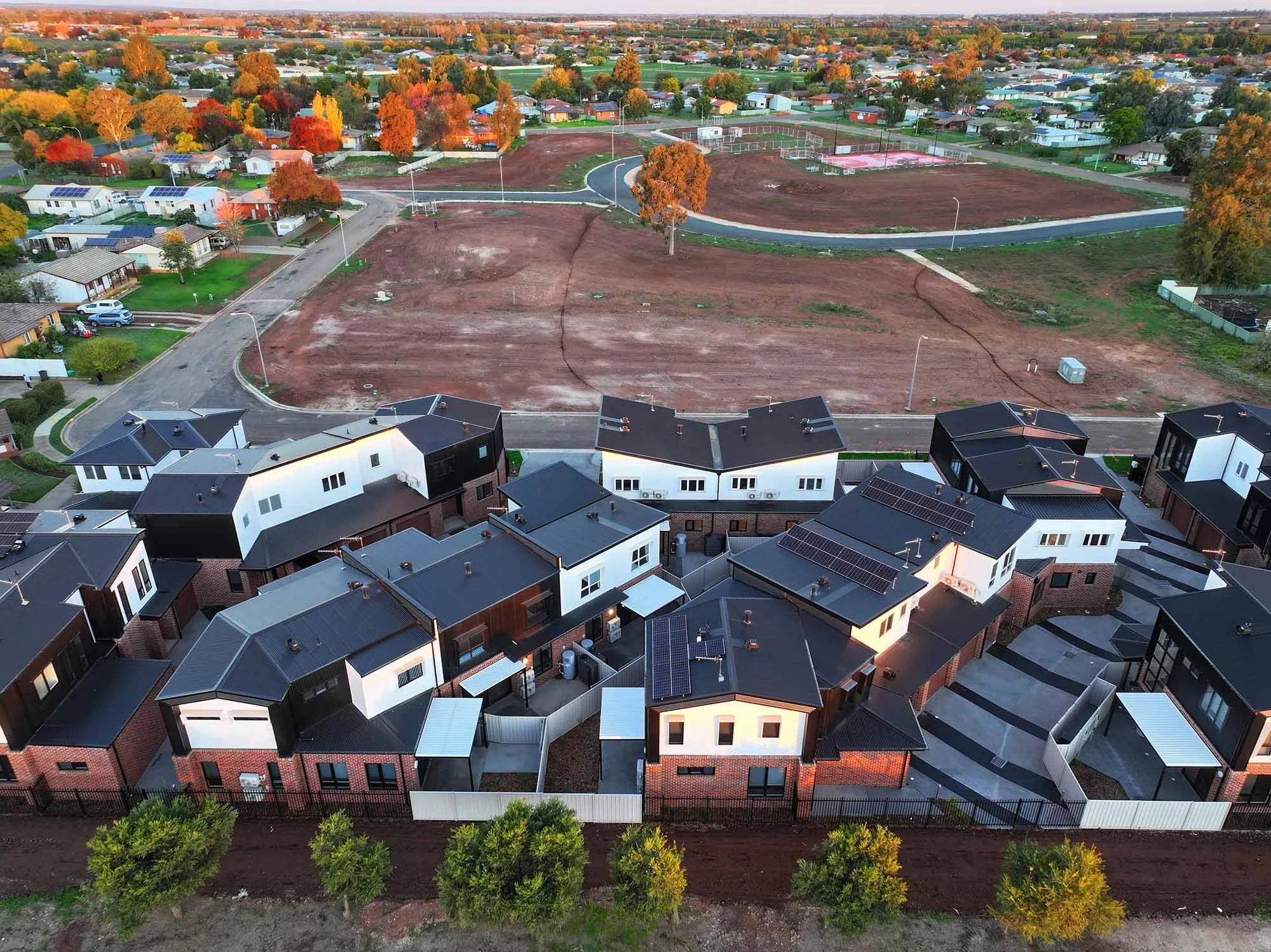 Aerial view of a neighborhood with new construction homes and an open lot being developed, surrounded by trees with fall foliage.