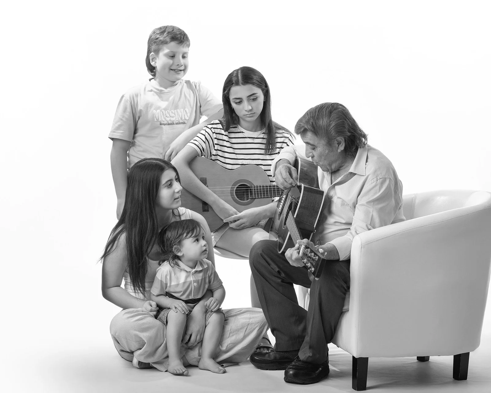 An elderly man playing acoustic guitar surrounded by four young girls, some holding guitars, in a studio with a white background.