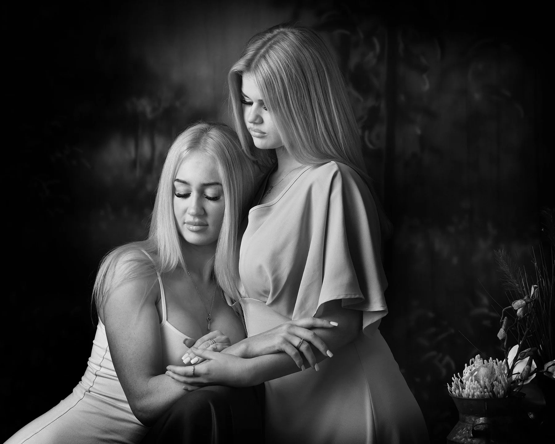 Black and white photo of two young women, one standing and one sitting, embracing each other with eyes closed, in front of a dark, blurred background with plants and flowers nearby.