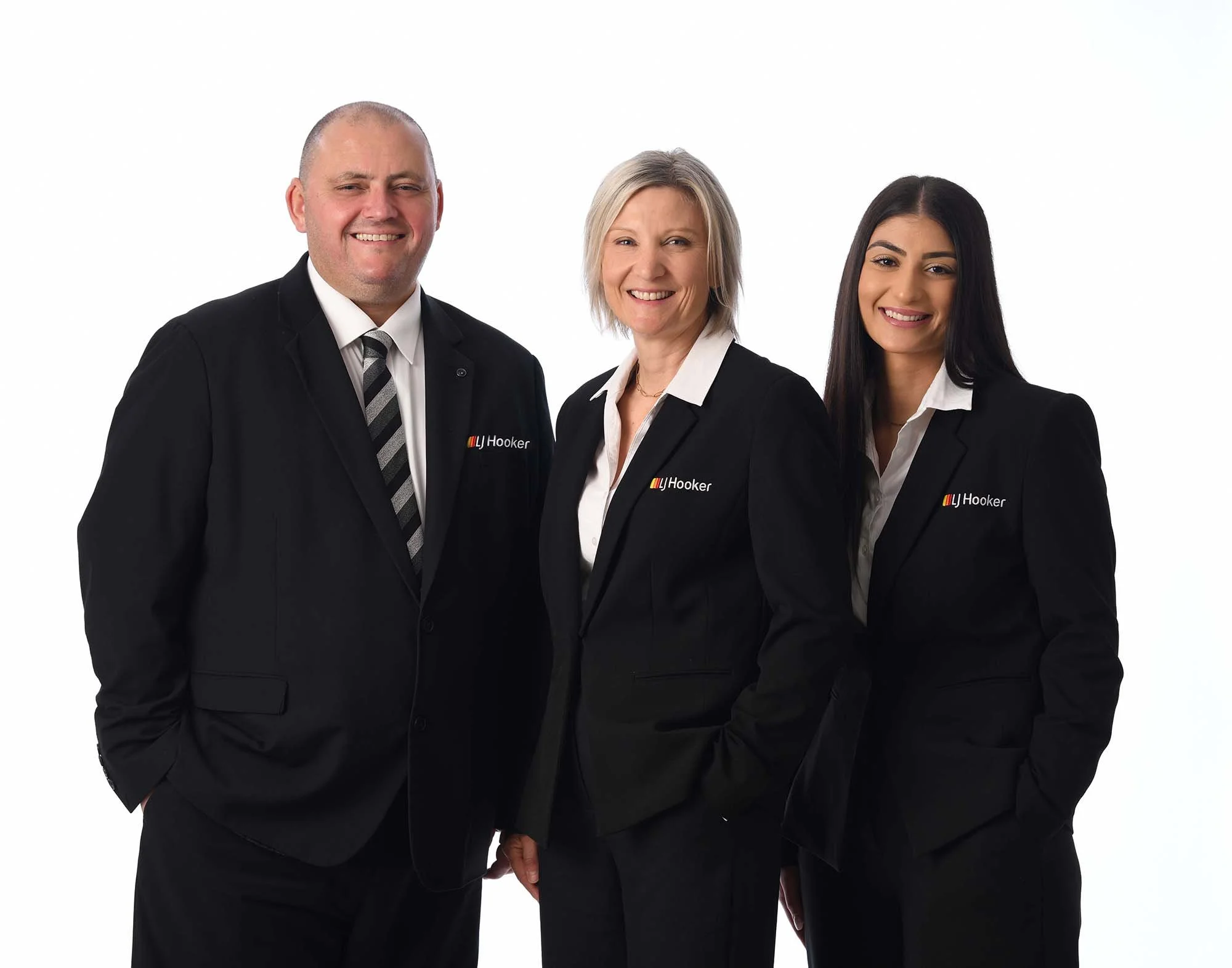 Three people dressed in professional black suits, two women and one man, standing against a plain white background, smiling at the camera.