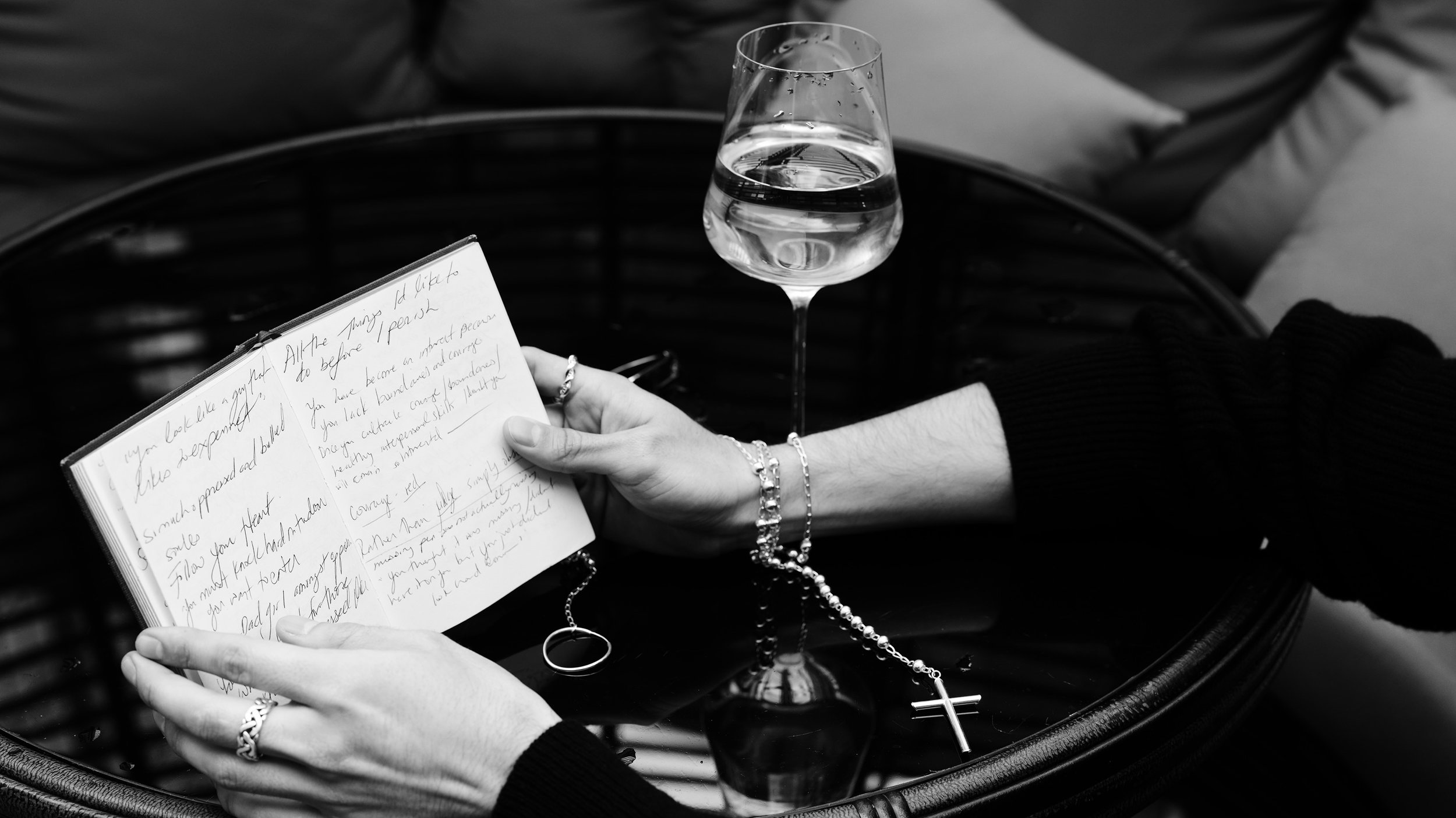 A person holding an open handwritten letter in one hand while sitting at a table. On the table, there is a glass of water and a pair of glasses. The person is wearing jewelry, including a ring, bracelet, and necklace with a cross pendant.