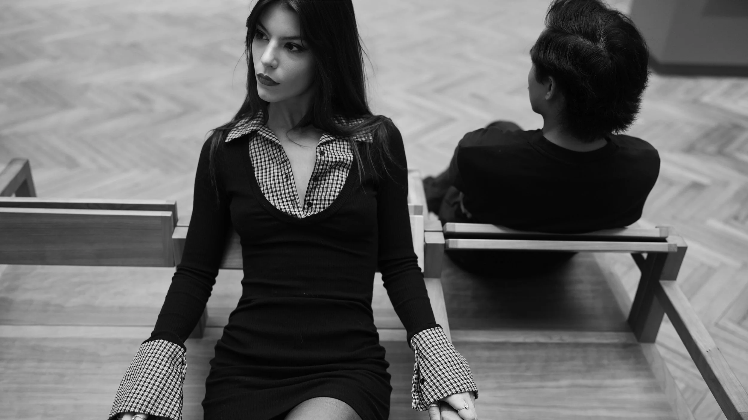 A woman with dark hair and lipstick sitting on a wooden bench, looking to the side, with a man with dark, wavy hair sitting nearby, facing away, in a modern indoor setting.