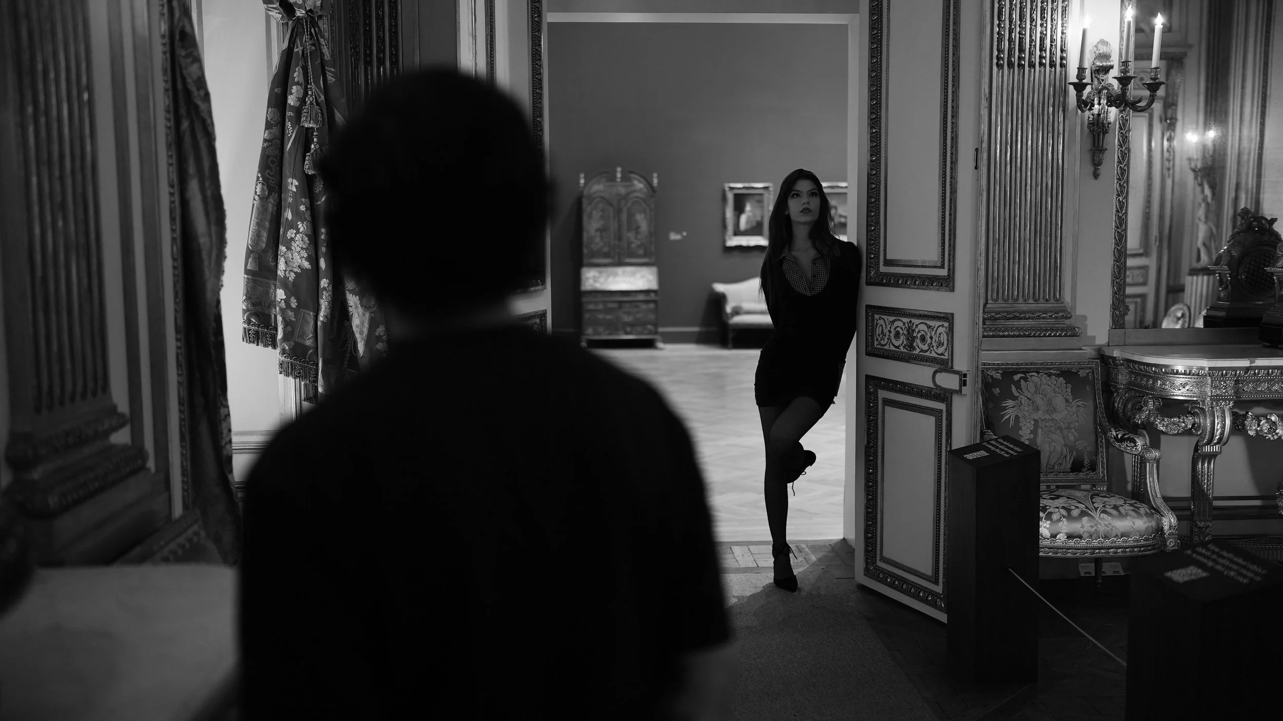 A woman standing in a museum gallery, leaning against a decorative doorframe, with ornate furnishings and artwork visible in the background. The image is in black and white.