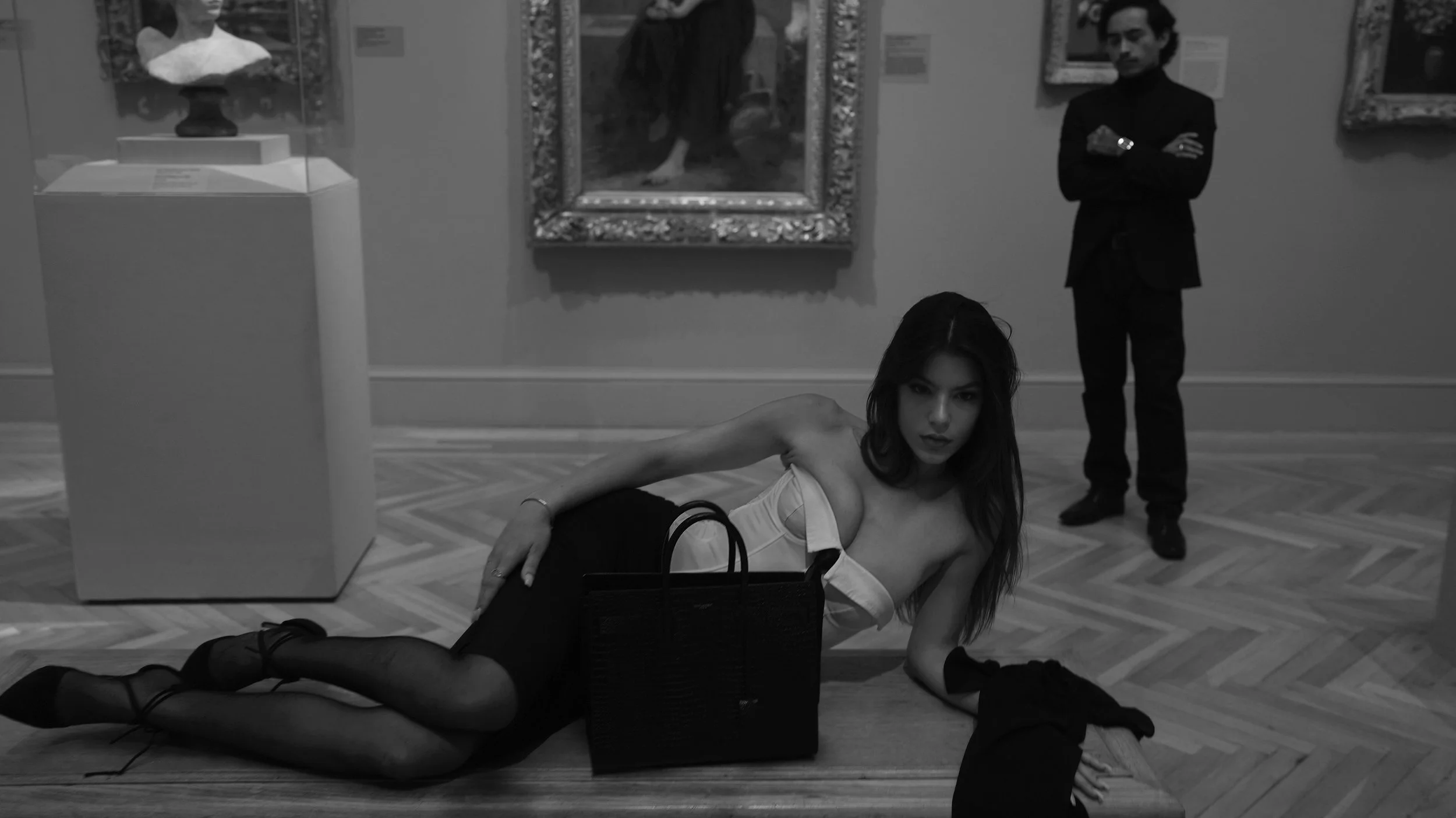 A woman lies on the floor of an art gallery, posing with her head on her arm and looking at the camera. She is wearing a white top, black stockings, and high heels, with a handbag on the floor beside her. In the background, a man stands with arms cro