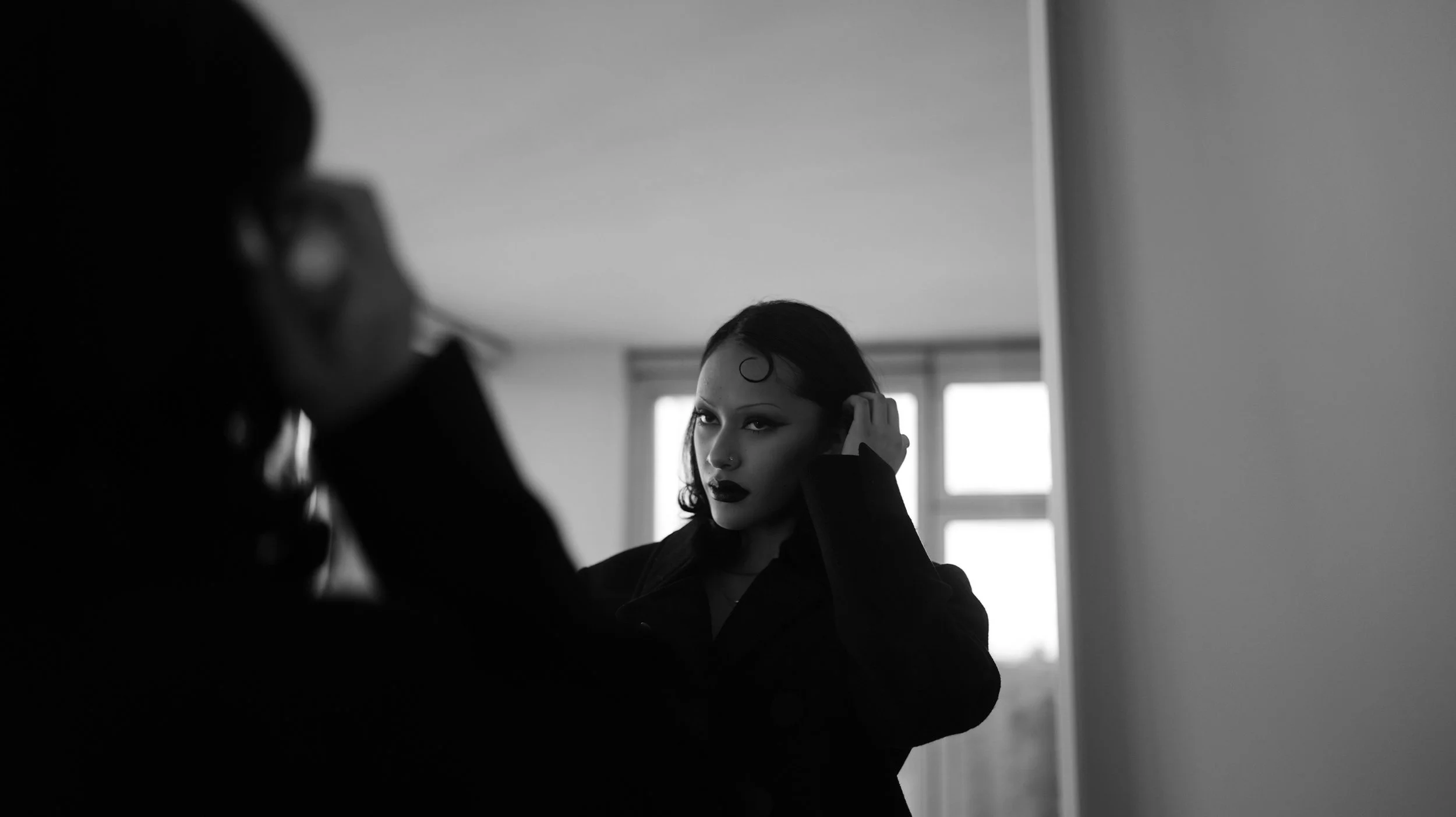 A woman with dark hair and makeup touching her ear while looking into a mirror, with a second person blurred in the foreground.