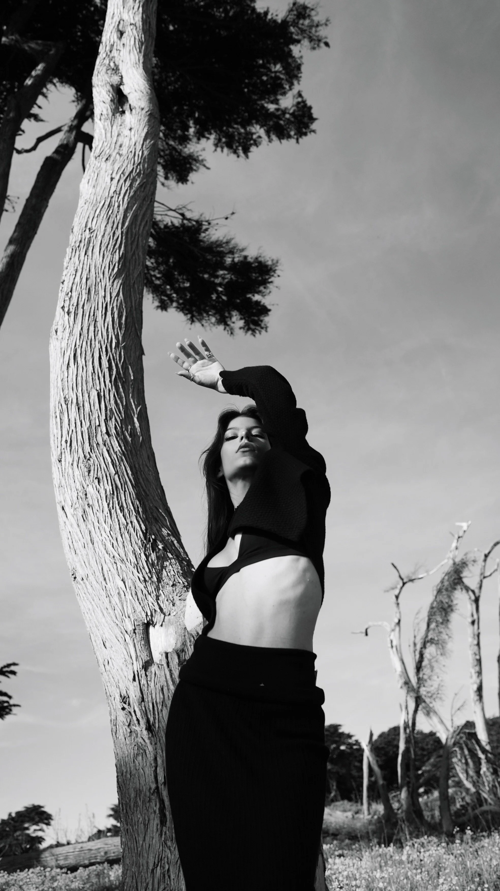 A black and white photograph of a woman standing outdoors near a large, textured tree trunk with her arm raised above her head. She is wearing a black crop top and a black skirt or pants, and has long hair and makeup with a confident expression. The 