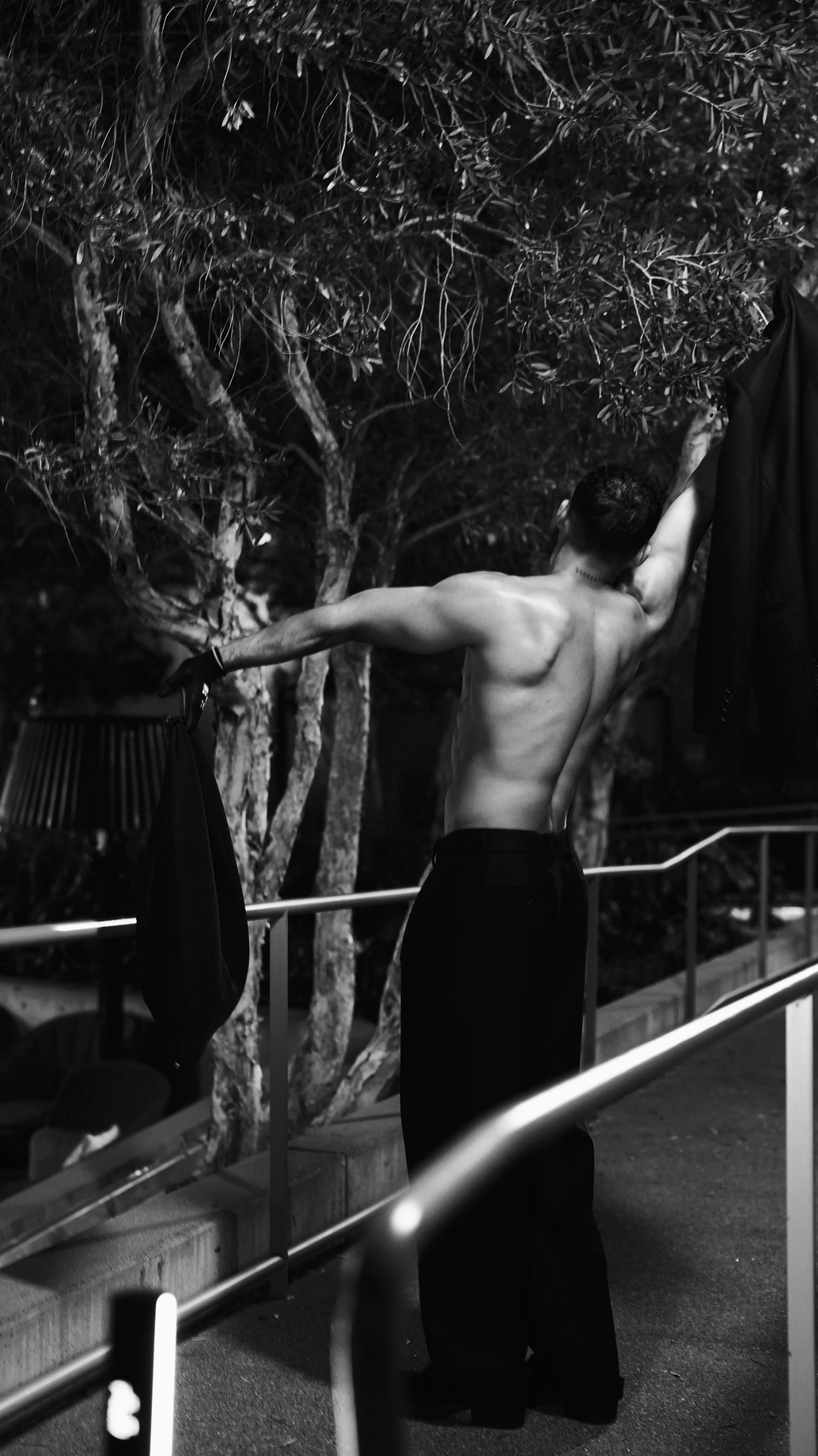 A shirtless man with short dark hair and tattoos on his neck is stretching his arms, holding a jacket in one hand in a park or outdoor area at night. Trees are in the background.