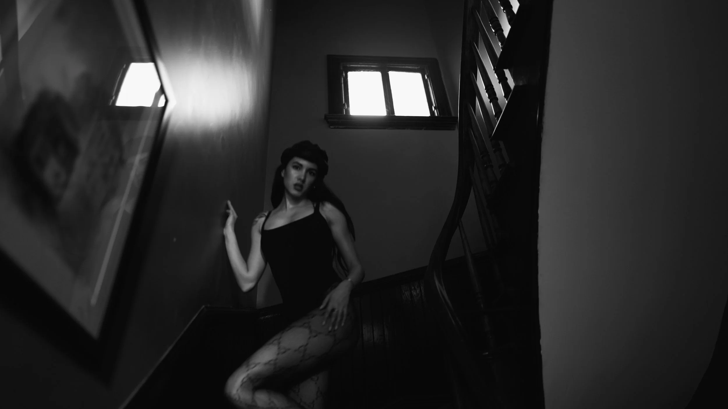 A black and white photo of a woman with dark hair, wearing a sleeveless dress, standing in a dimly lit stairwell next to a window.