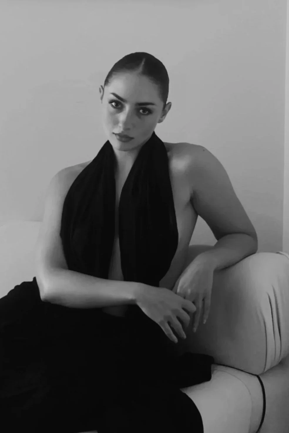 A black-and-white photo of a woman with slicked-back hair, sitting shirtless on a beige armchair, wearing dark pants and a scarf around her neck, looking directly at the camera with a neutral expression.